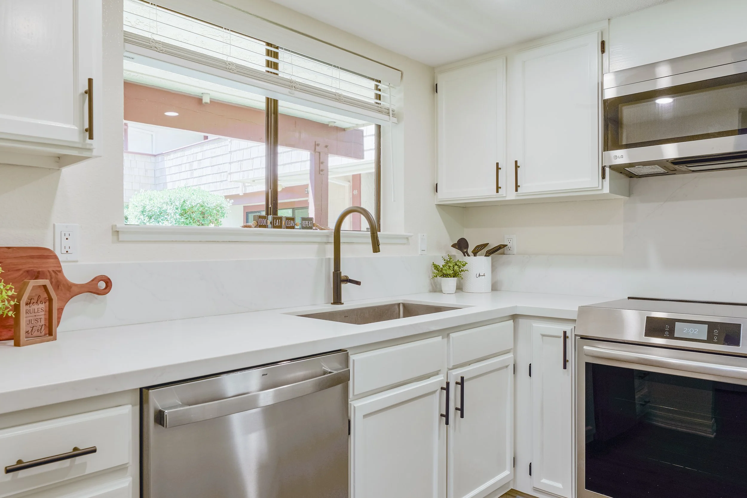 A modern kitchen with white cabinets, stainless steel dishwasher, oven, and a window above the sink. On the windowsill, there are decorative blocks with words, plants, and window blinds.