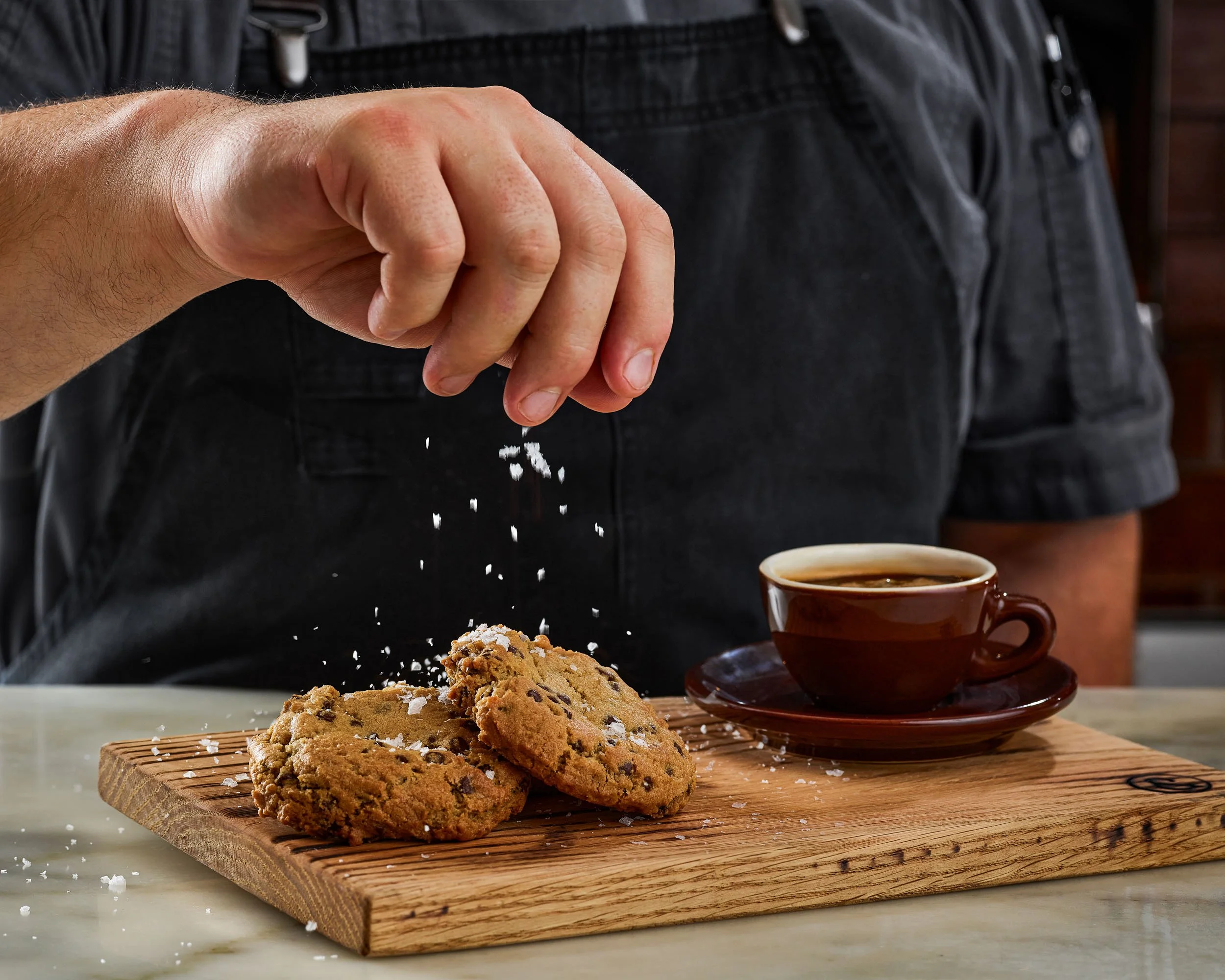 Close-up of a person's hand sprinkling salt on freshly baked chocolate chip cookies placed on a wooden cutting board with two brown coffee cups in the background.