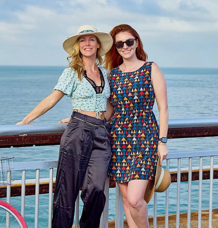 Two women smiling and posing on a seaside pier, with ocean and cloudy sky in the background. One woman wears a wide-brimmed hat, sunglasses, a cropped blouse, and loose pants; the other wears sunglasses, a colorful geometric dress, and holds a hat.