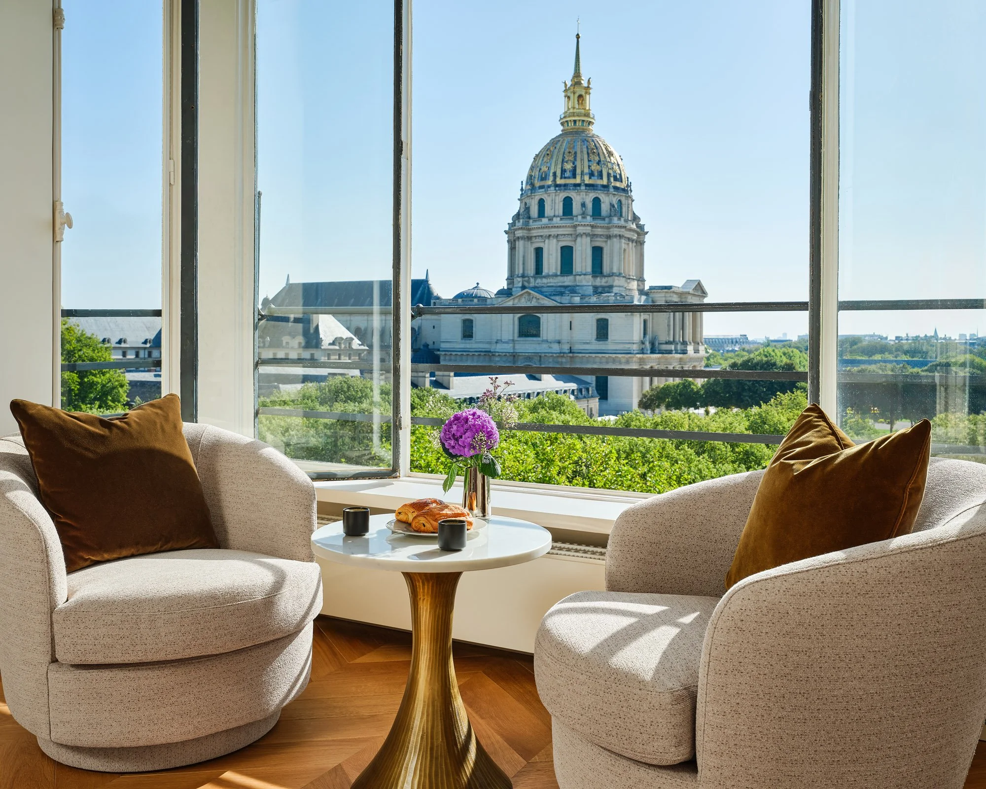 Living room with large open window showing a historic building with a domed roof and tower, greenery outside, and furniture including a beige sofa with brown pillows, a white round table with a vase of purple flowers, a croissant, and two black cups.