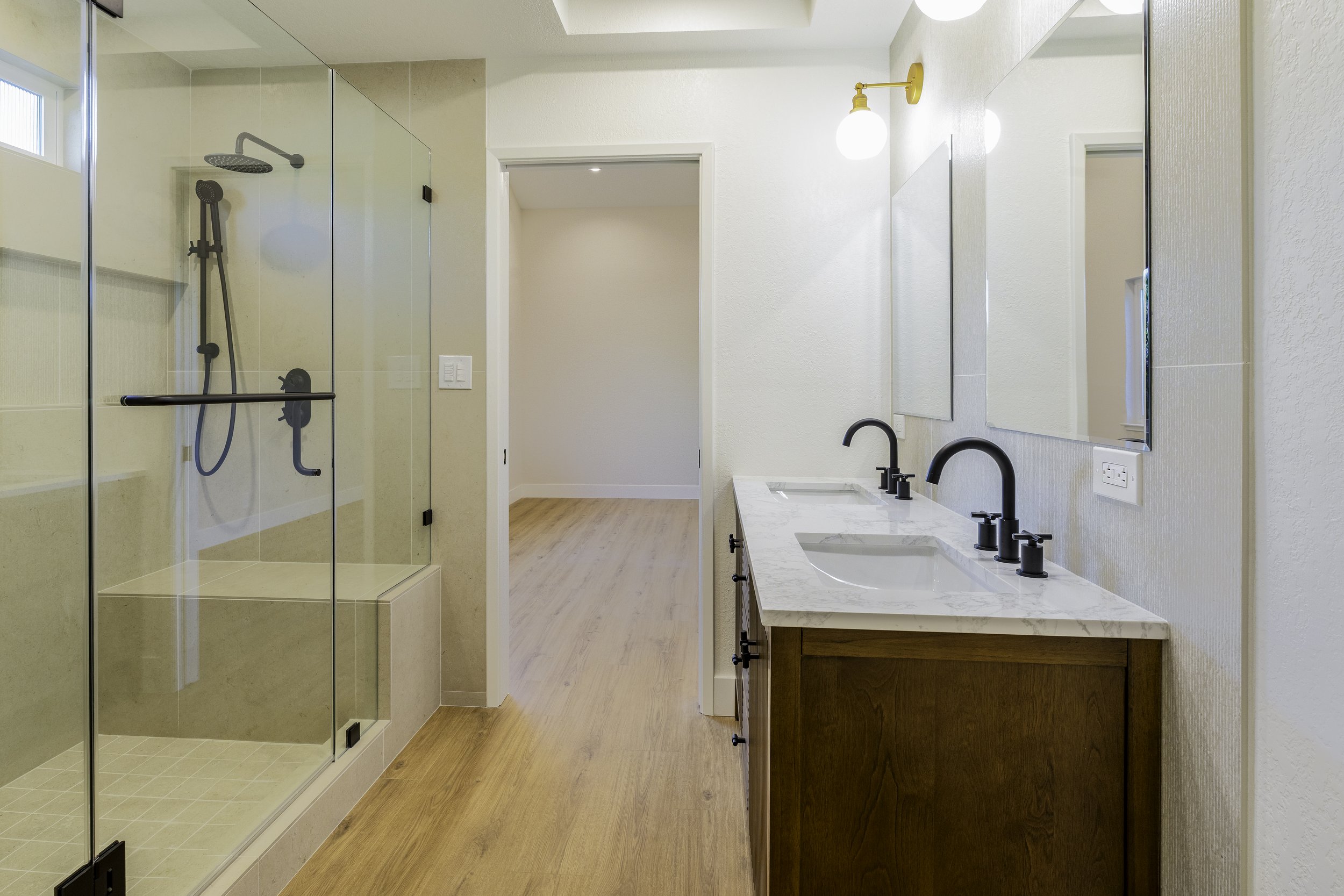 A modern bathroom with a walk-in shower with glass doors on the left, two sinks with black faucets and a wooden vanity on the right, a large mirror, white walls, and wood flooring.
