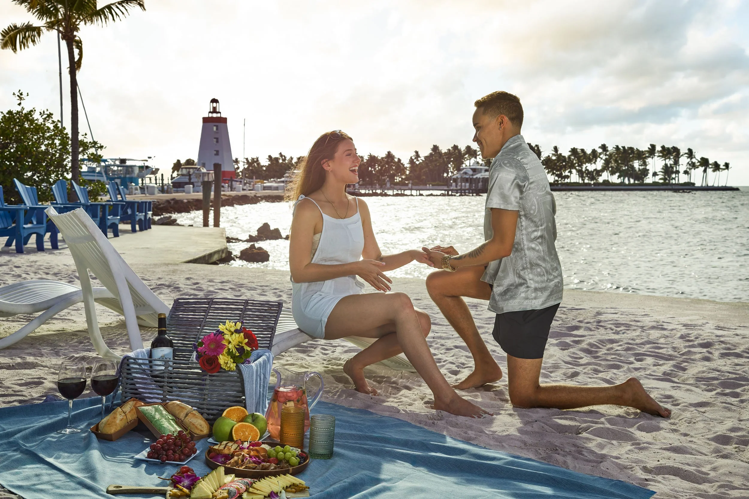 A couple is having a romantic proposal on a beach, with a picnic setup including fruits, wine, and flowers, as one man kneels holding the woman's hand. The background features a lighthouse, boats, and a palm-lined shoreline during sunset.