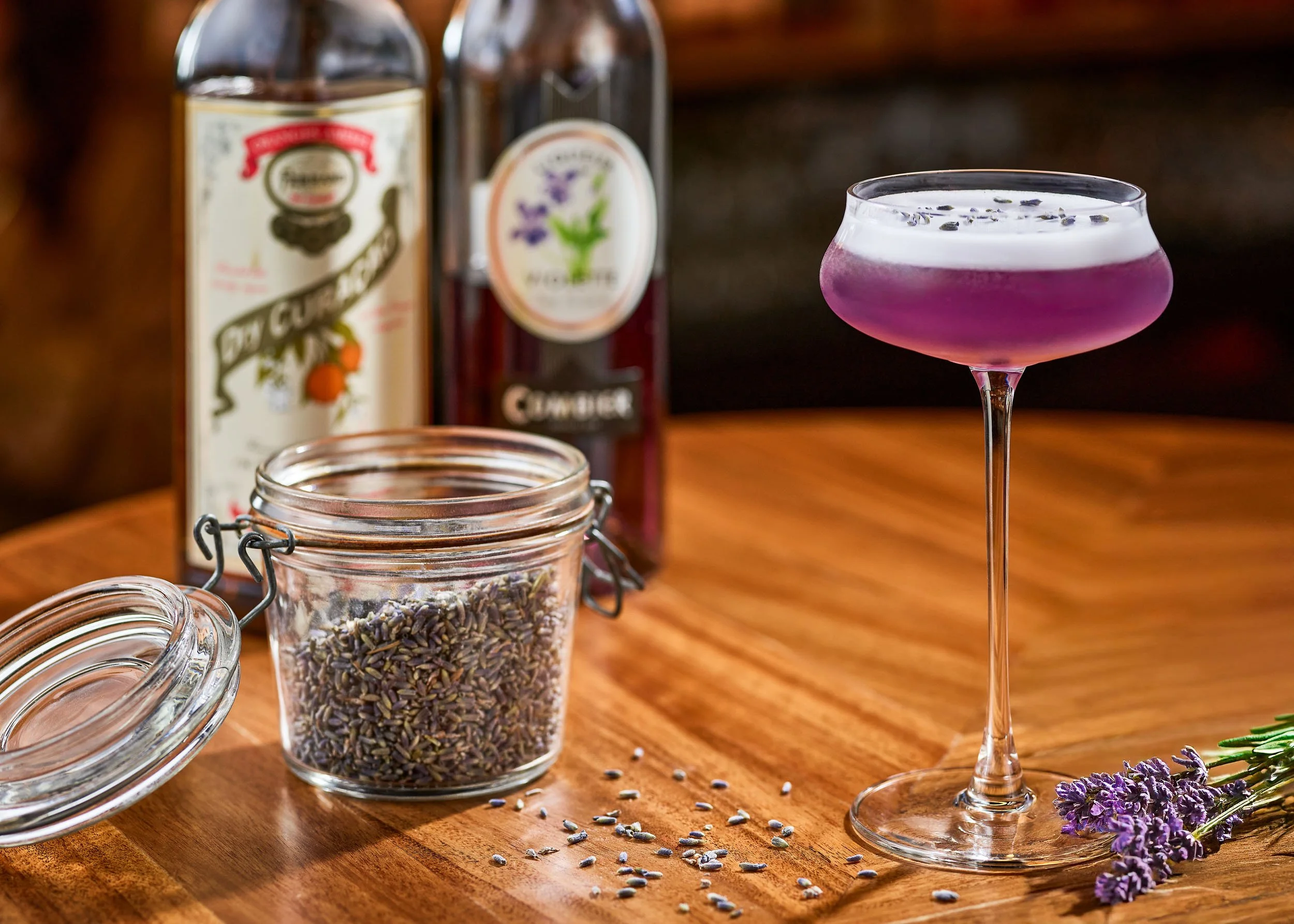 A cocktail glass with a pinkish-purple drink garnished with lavender flowers, placed on a wooden surface with scattered lavender seeds. Behind are bottles of flavored syrup and a jar of dried lavender buds.