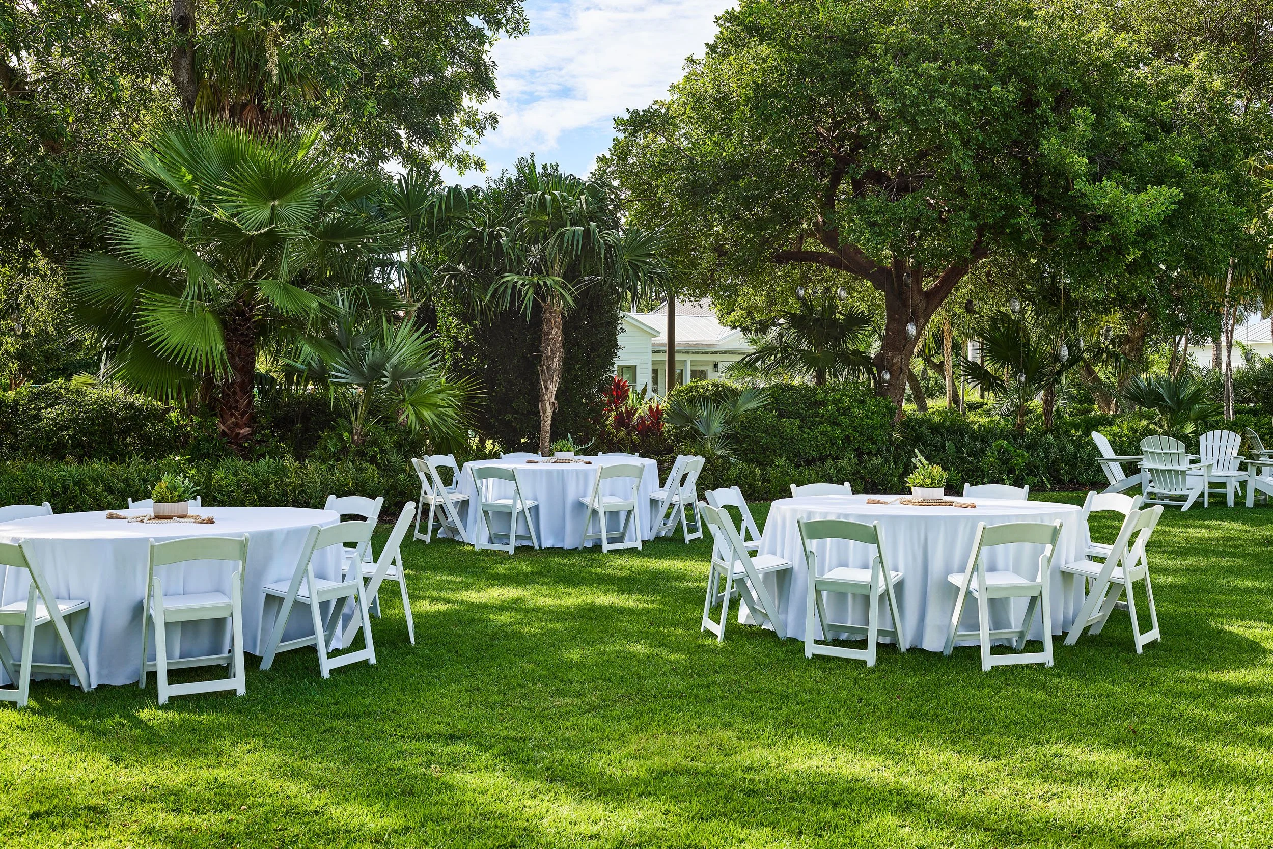 Outdoor event setup with round tables covered in white tablecloths, white folding chairs, and small potted plants, surrounded by lush green trees and foliage under a partly cloudy sky.