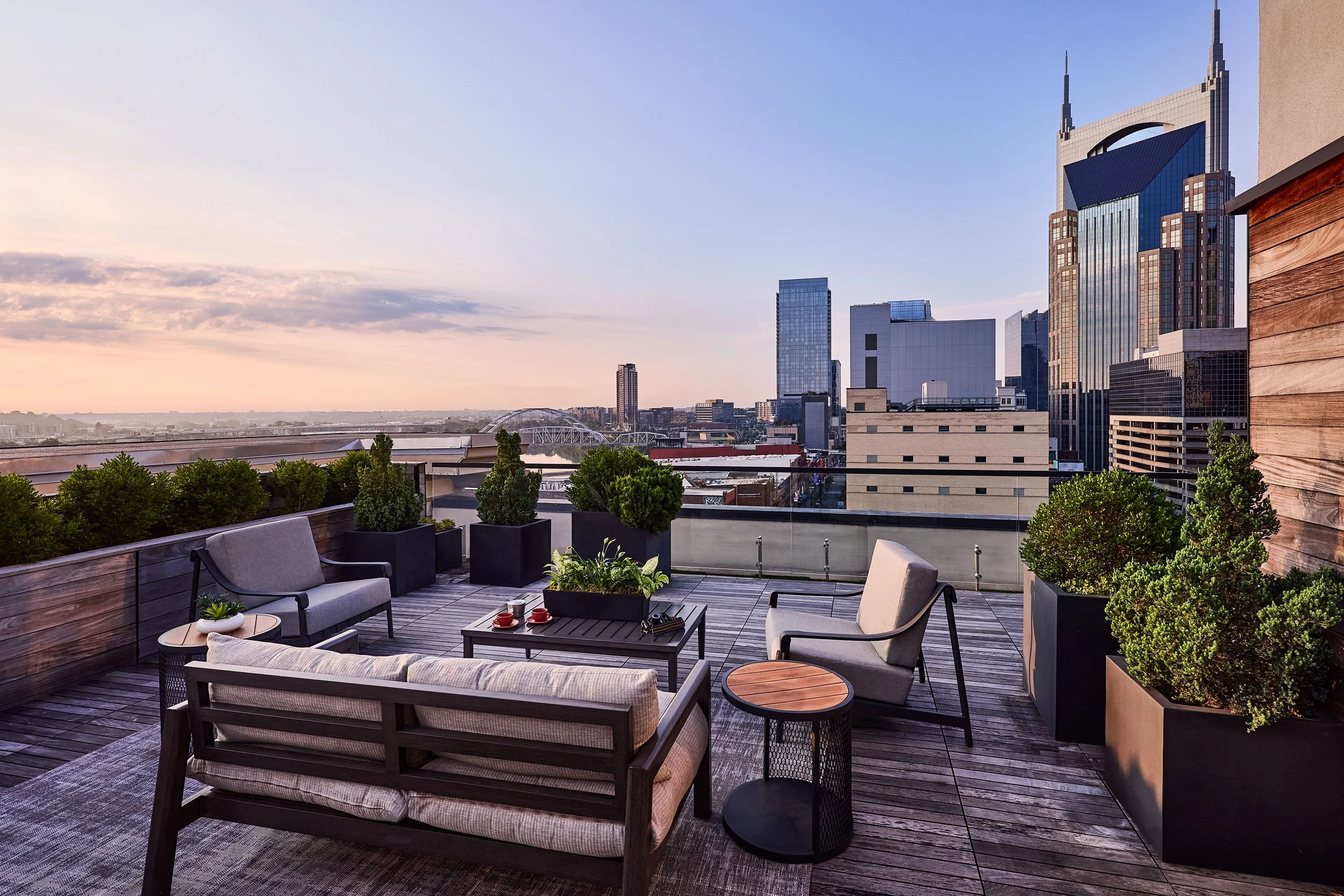 City skyline view from a rooftop patio, with modern high-rise buildings, trees, and a bridge against a sunset sky.