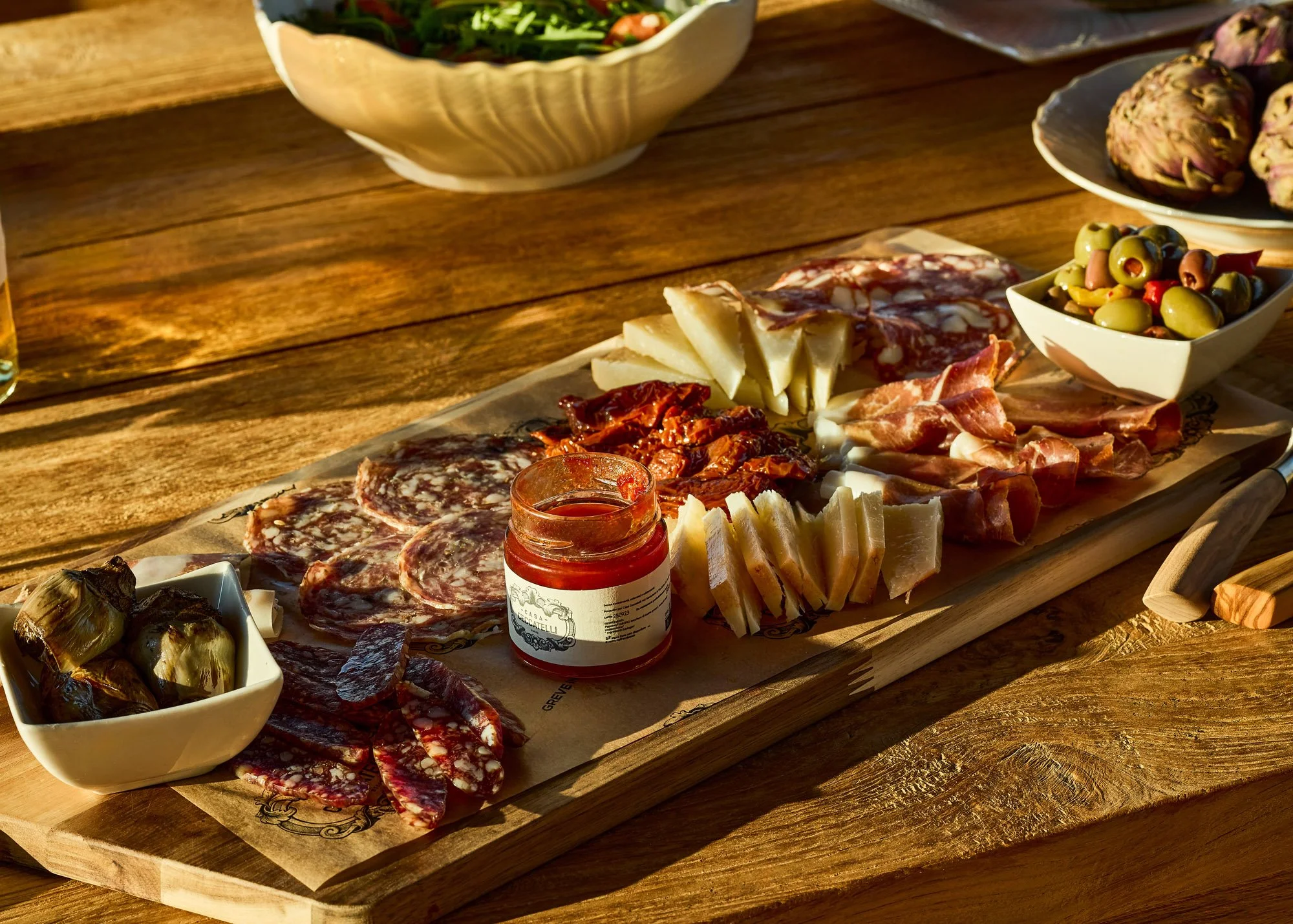 A charcuterie board featuring sliced cured meats, cheeses, green olives, roasted artichokes, and a jar of tomato sauce, sitting on a wooden table surrounded by salads and plates.