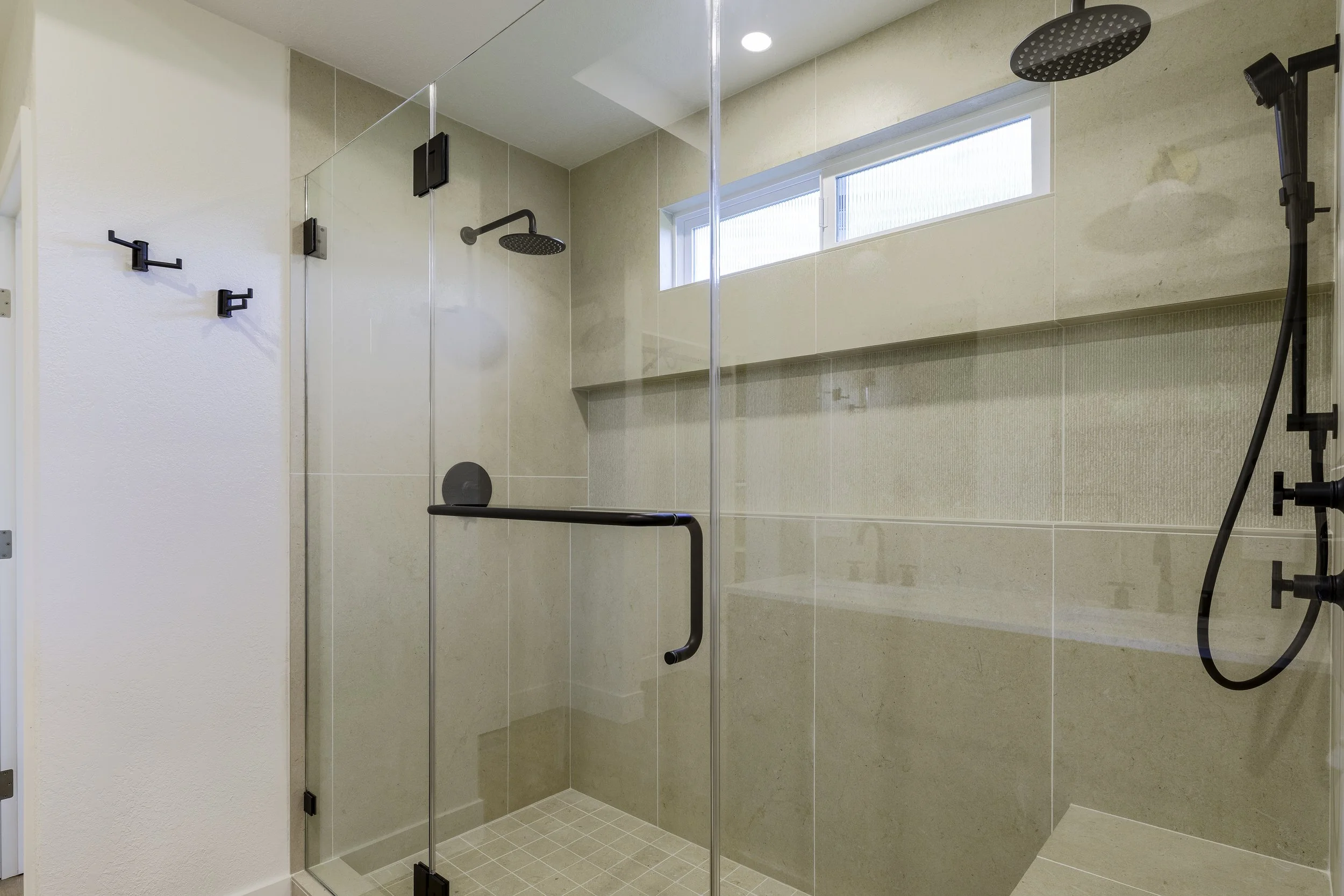 A modern walk-in shower with clear glass enclosure, black fixtures, rainfall showerhead, handheld shower, beige tiled walls, and a narrow window for natural light.
