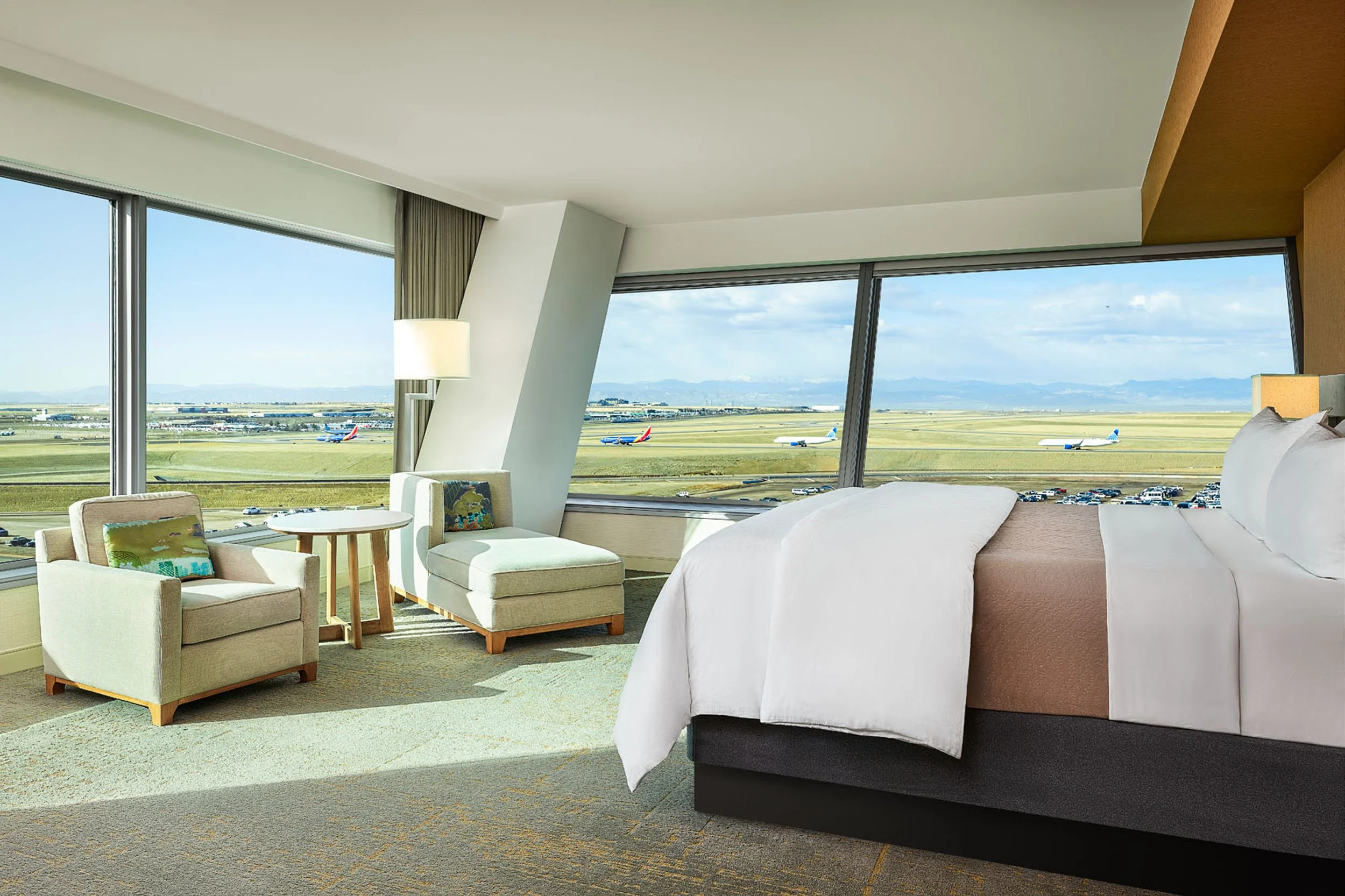 A hotel room with large windows showing planes on the runway outside, a bed with white linens, a purple headboard, and seating area with beige chairs and a small round table.