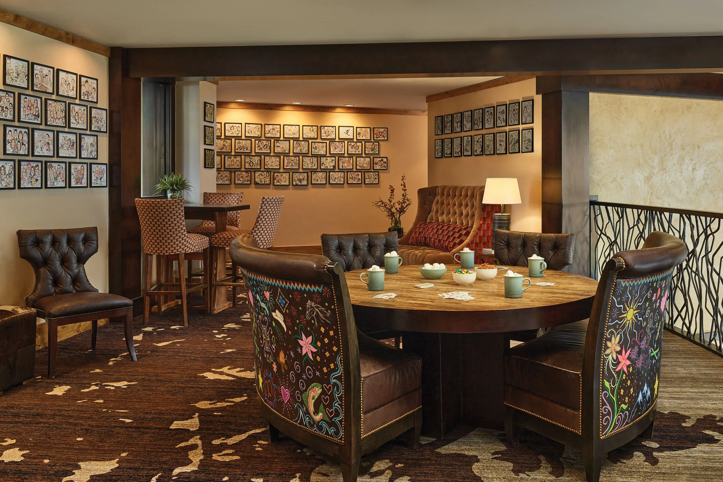A cozy indoor room with a round table set for a game or snack, surrounded by various cushioned chairs. The walls are decorated with framed artwork and a plant, and there is a softly lit corner with a lamp, creating a warm atmosphere.