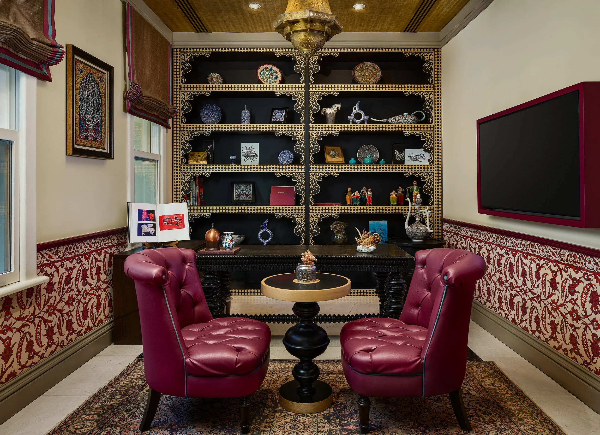 A cozy room with a black shelving unit decorated with ornate plates, vases, and artwork, flanked by windows with red and pink curtains, and four pink armchairs surrounding a round wooden table with decorative items.