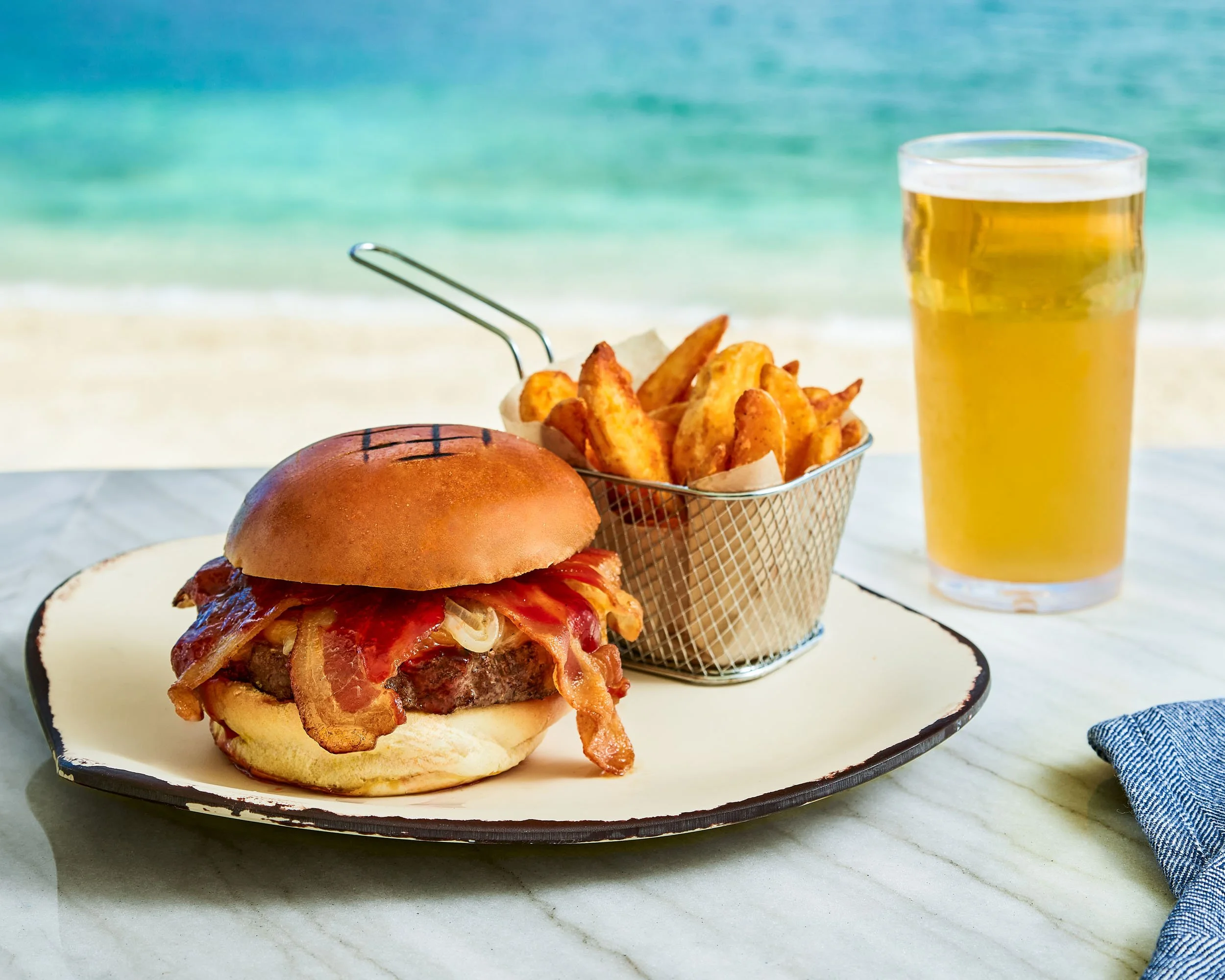 A cheeseburger with bacon on a white plate, a basket of French fries, and a glass of beer on a table near the beach with ocean in the background.