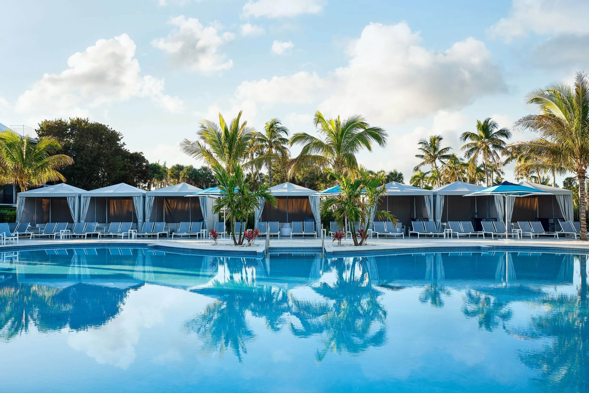 A tropical swimming pool area with lounge chairs and cabanas, surrounded by palm trees under a partly cloudy sky, with the pool's reflection visible in the water.