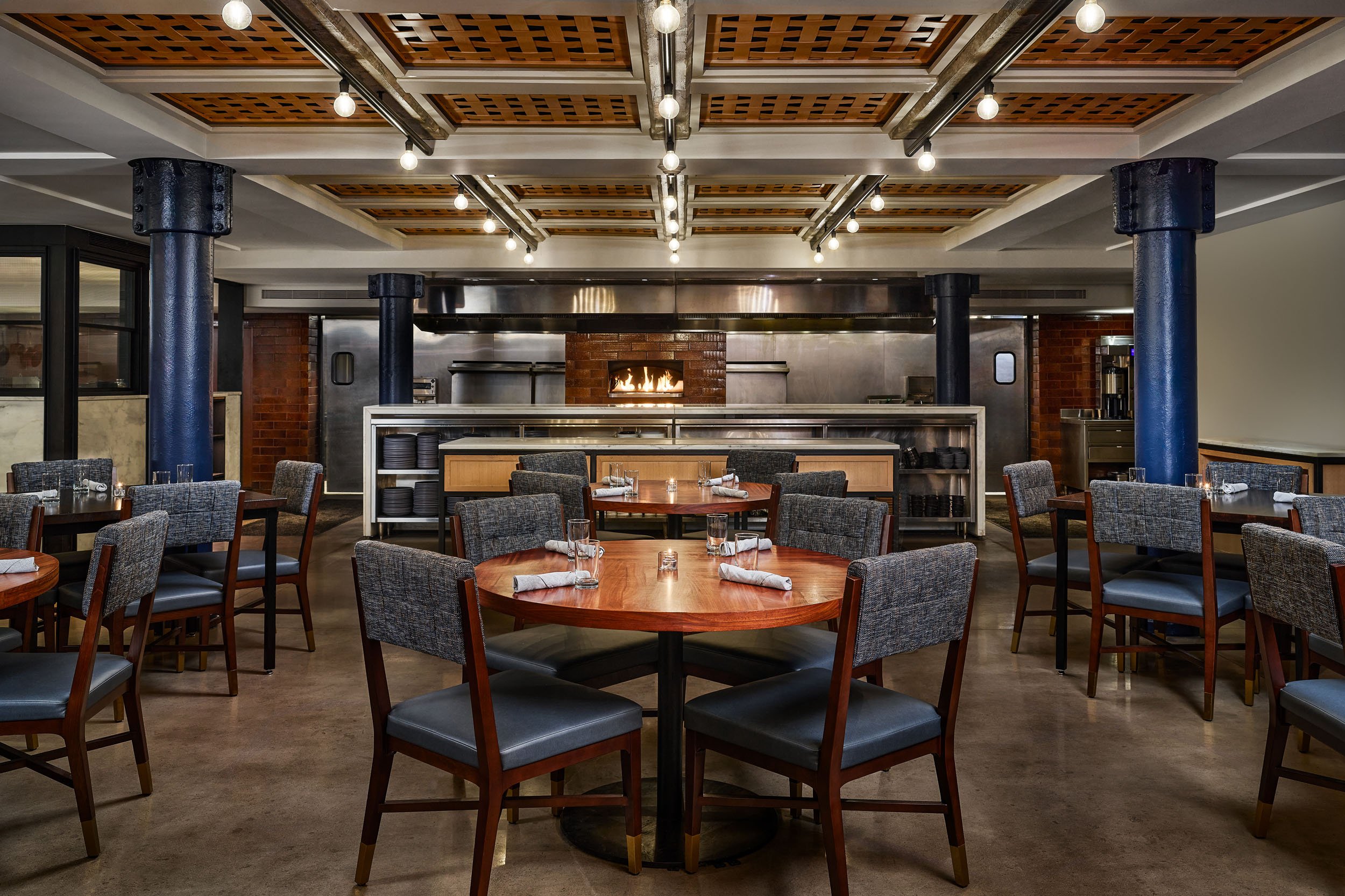 Interior of a restaurant with round tables set with napkins, glasses, and cutlery. The background features a fireplace, black columns, wooden ceiling panels, and modern lighting fixtures.