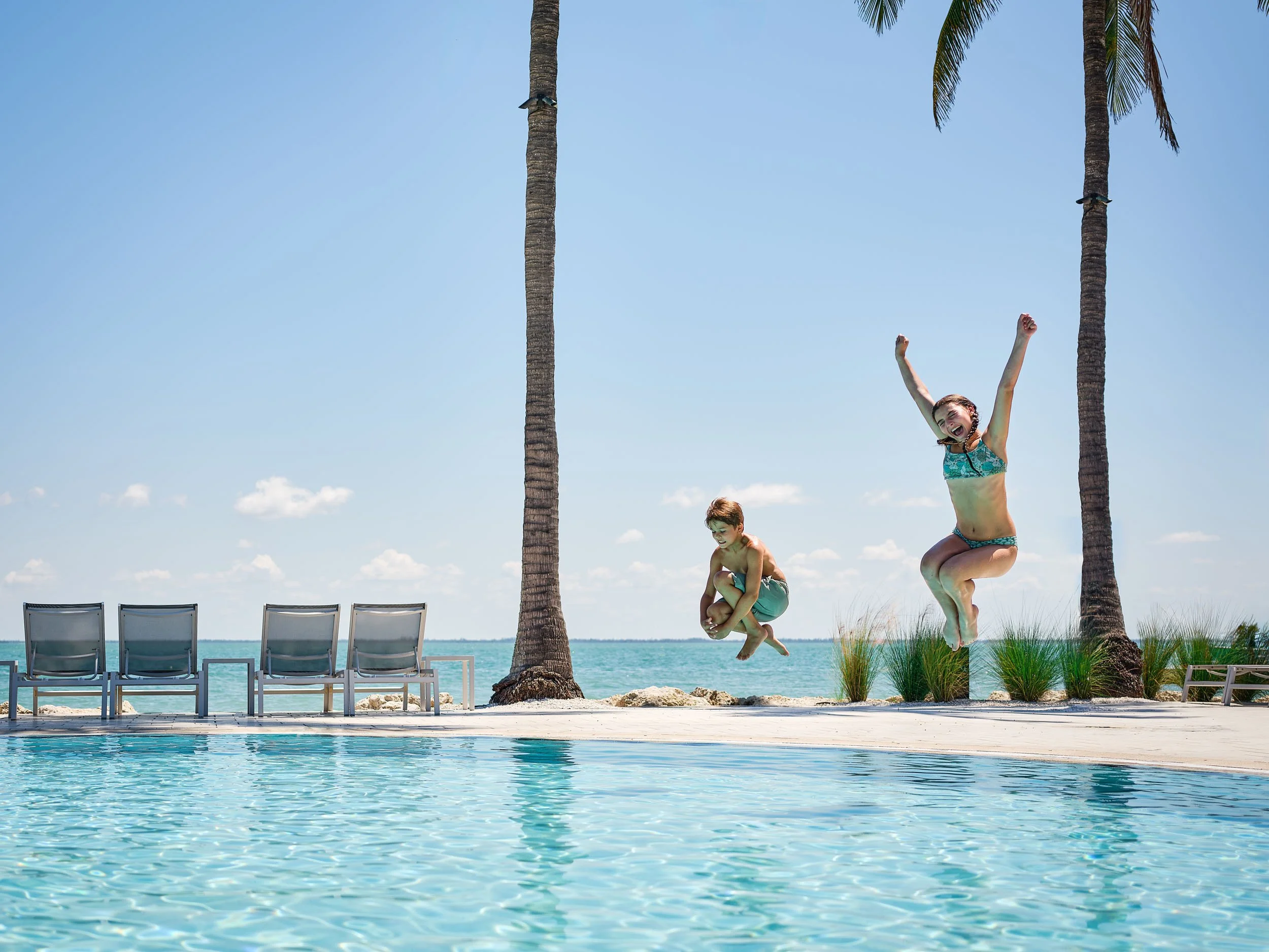 A girl in a blue swimsuit jumping into a swimming pool, with a boy crouching nearby, a row of chairs, palm trees, and the ocean in the background on a sunny day.