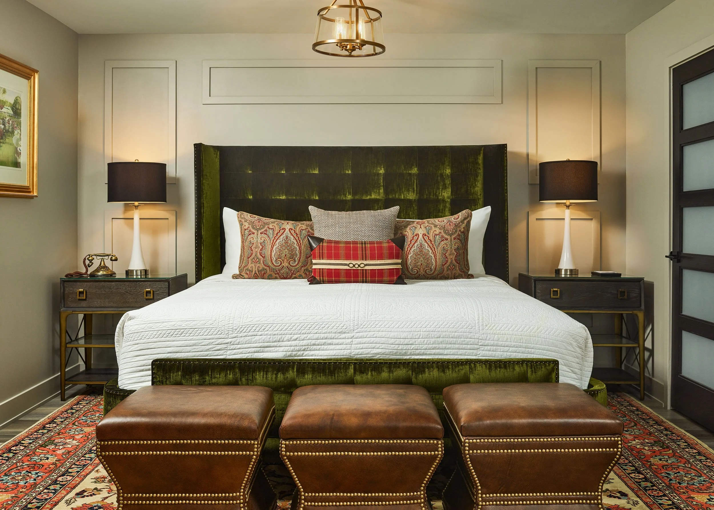 A bedroom with a green velvet upholstered headboard, a white bedspread, multiple decorative pillows, two matching black lamps on nightstands, leather ottomans at the foot of the bed, framed artwork on the walls, a ceiling light fixture, and a pattern