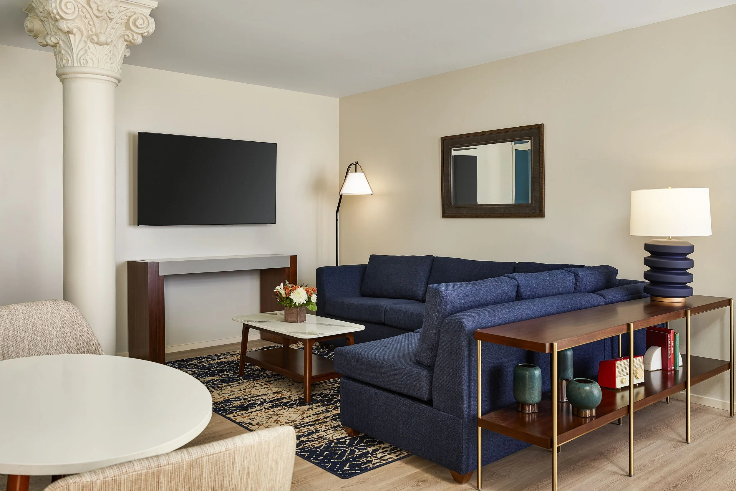 Living room with beige walls, a mounted flat-screen TV, a navy blue sofa, a wooden side table with green vases, a wall mirror, and a modern floor lamp.