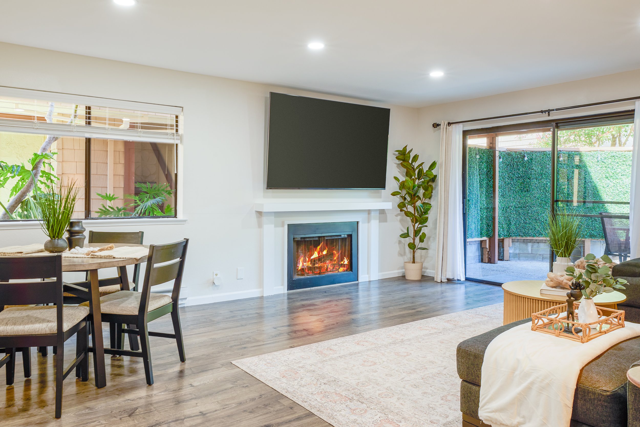 A living room with a fireplace, a wall-mounted TV, large windows with curtains, and a patio door opening to an outdoor space with greenery.