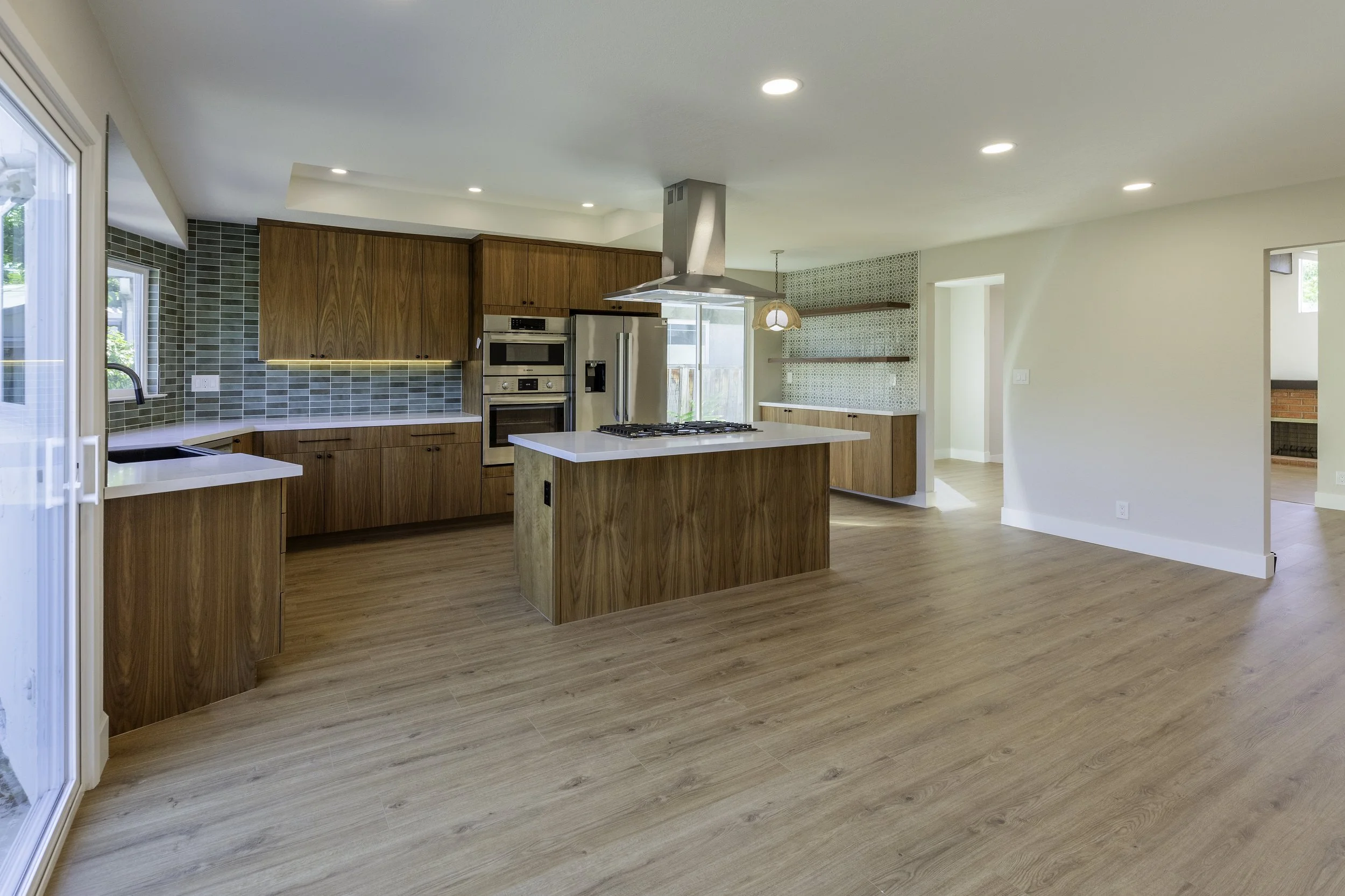 Open-concept kitchen with wooden cabinets, a kitchen island with a cooktop, stainless steel appliances, green tiled backsplash, and large windows for natural light.