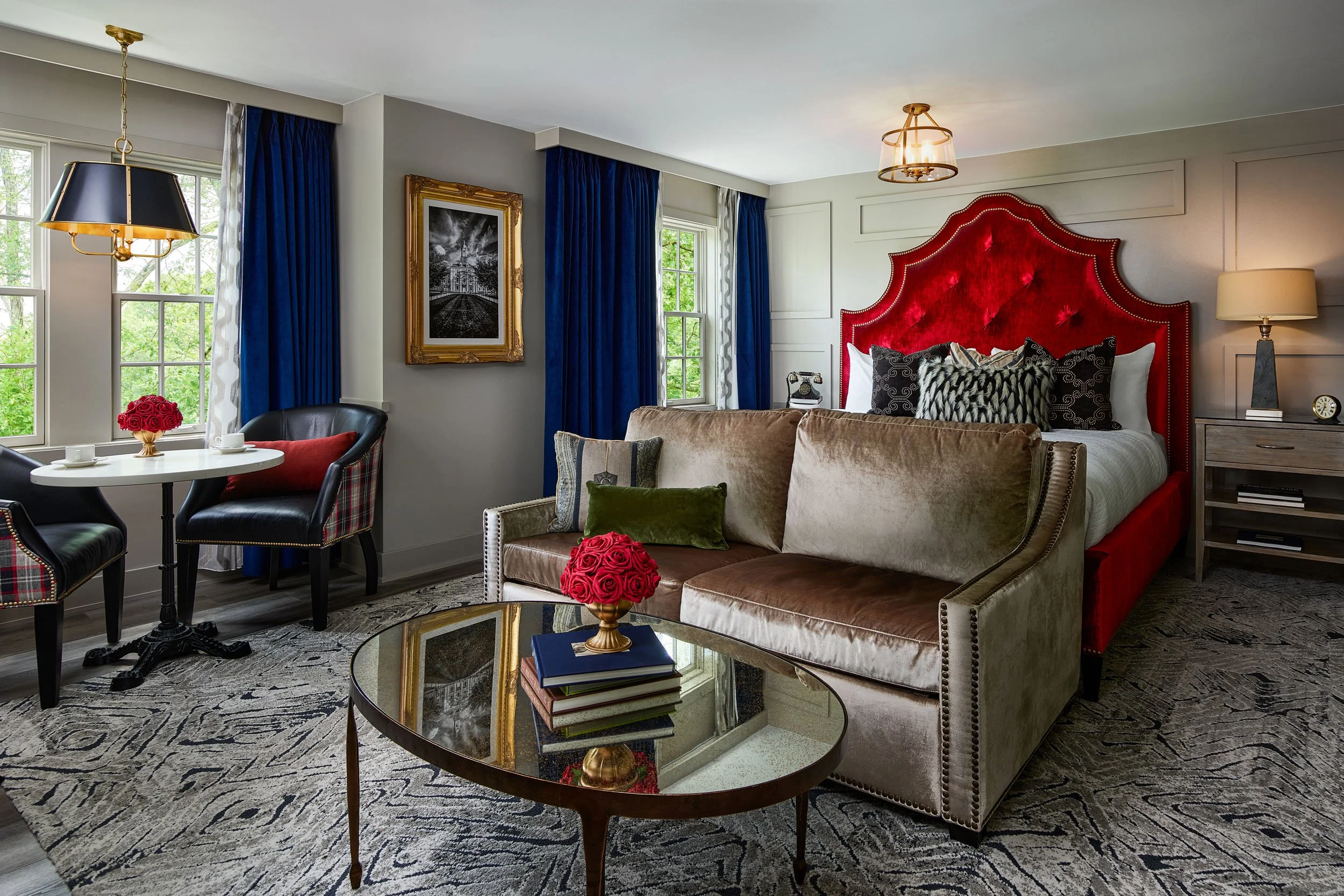 Living room with a large red upholstered headboard, velvet and patterned cushions, side table with lamp, window with blue curtains, round glass coffee table, and a seating area with chairs and flowers.