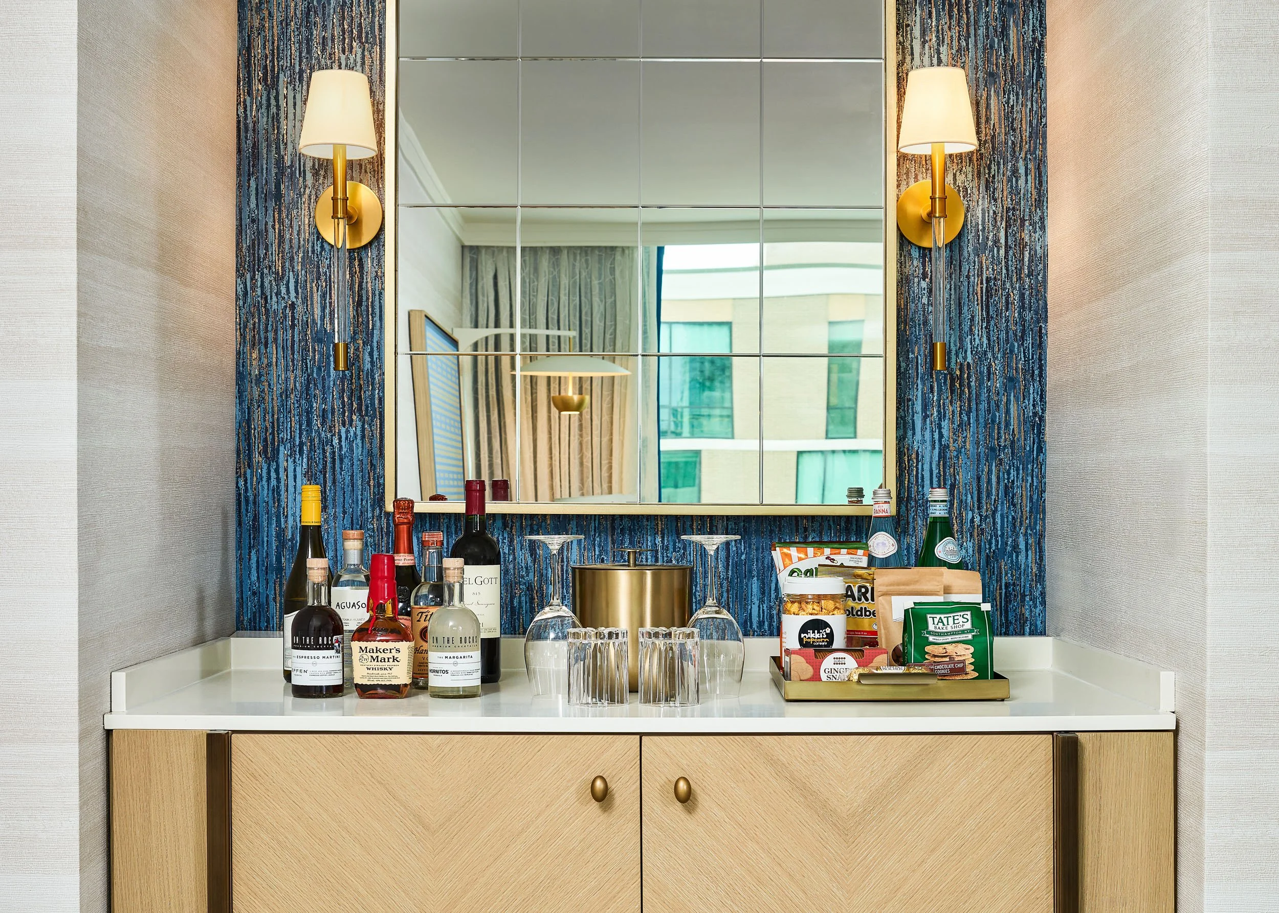 A bar or kitchen counter with several bottles of alcohol, glasses, snacks, and beverage bottles, with a large mirror and wall sconces with yellow shades behind. The reflection in the mirror shows a window and part of a bed or couch with curtains.