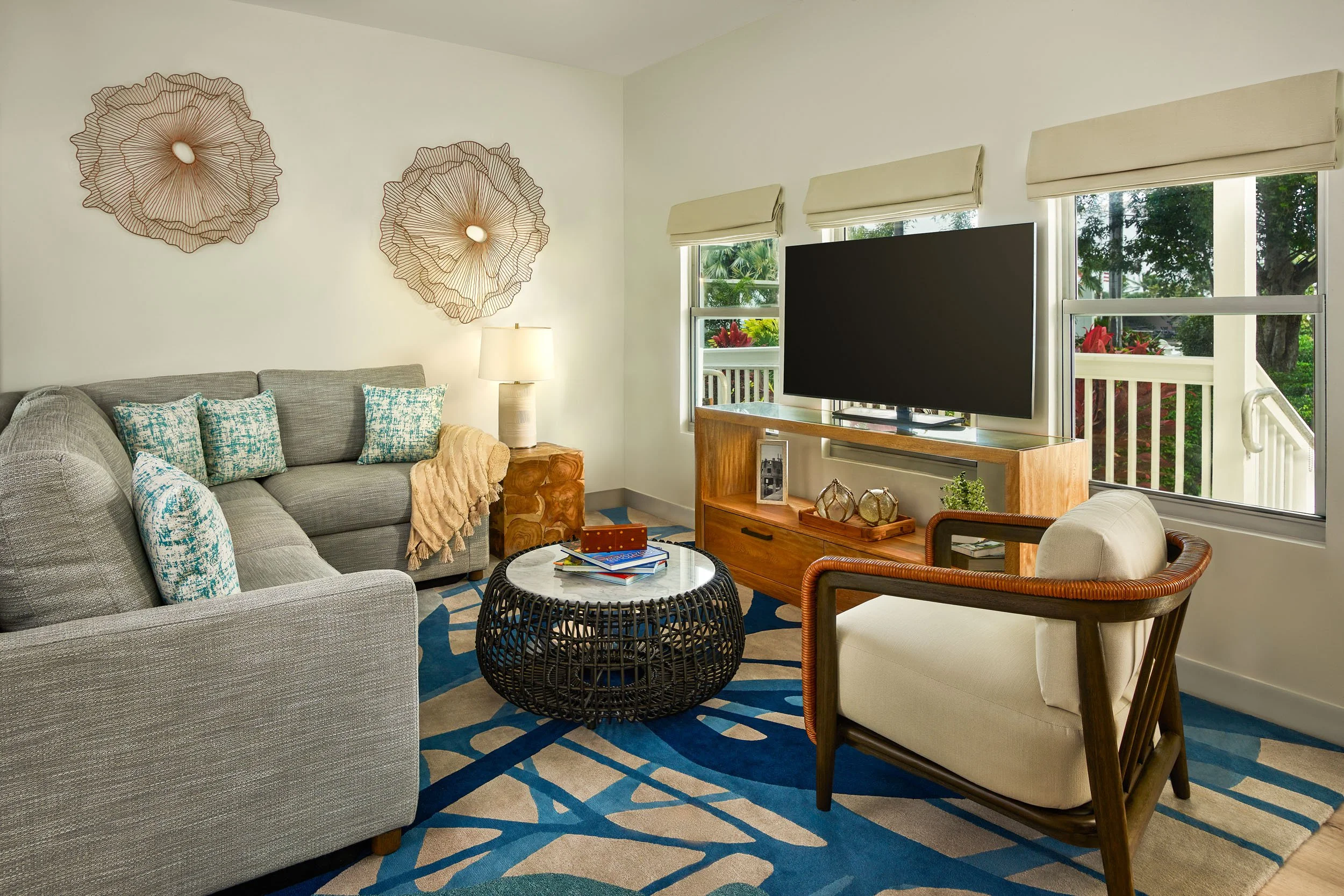 Living room with a grey sectional sofa, patterned pillows, wooden side table, and a black wicker coffee table. A wall-mounted flat-screen TV is on a wooden stand near three windows with beige blinds. Decor includes artwork, a table lamp, and plants v