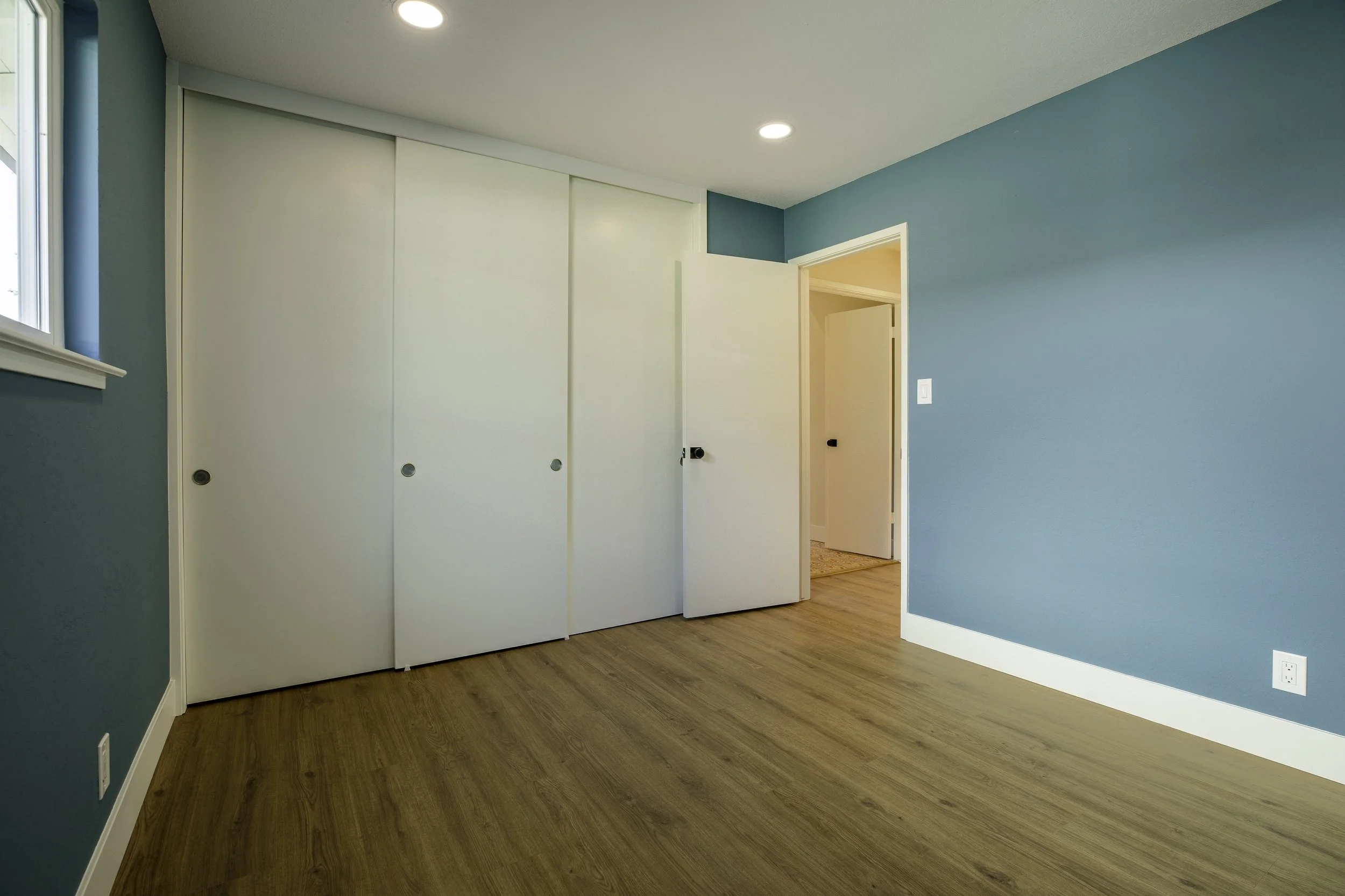 Room with teal walls, white closet doors, a window, wood flooring, and a partially open door leading to another room.