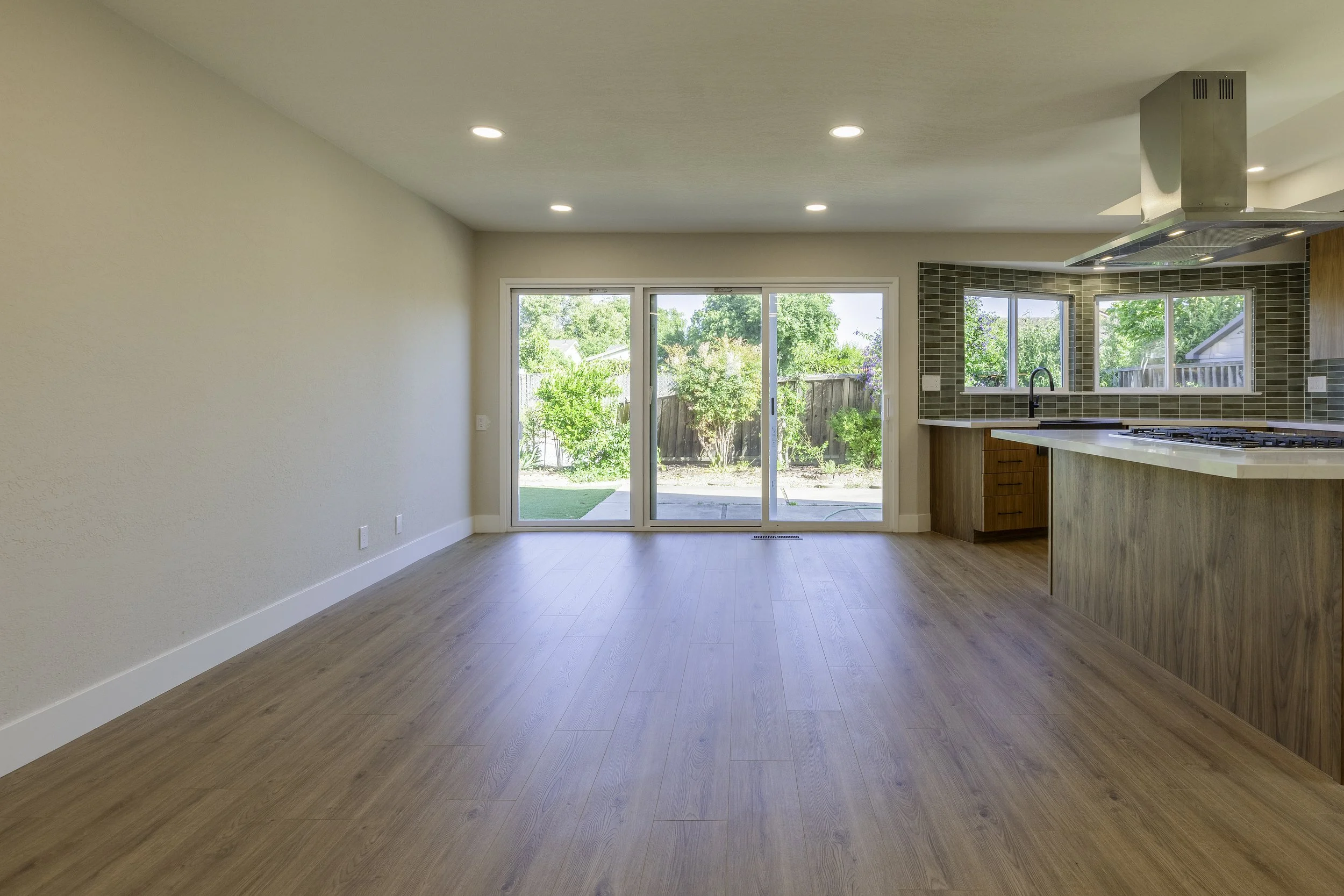 Empty living room with sliding glass doors leading to a backyard with trees and a wooden fence. Kitchen with windows, wooden cabinets, and a stove hood. Hardwood floors and recessed ceiling lights.