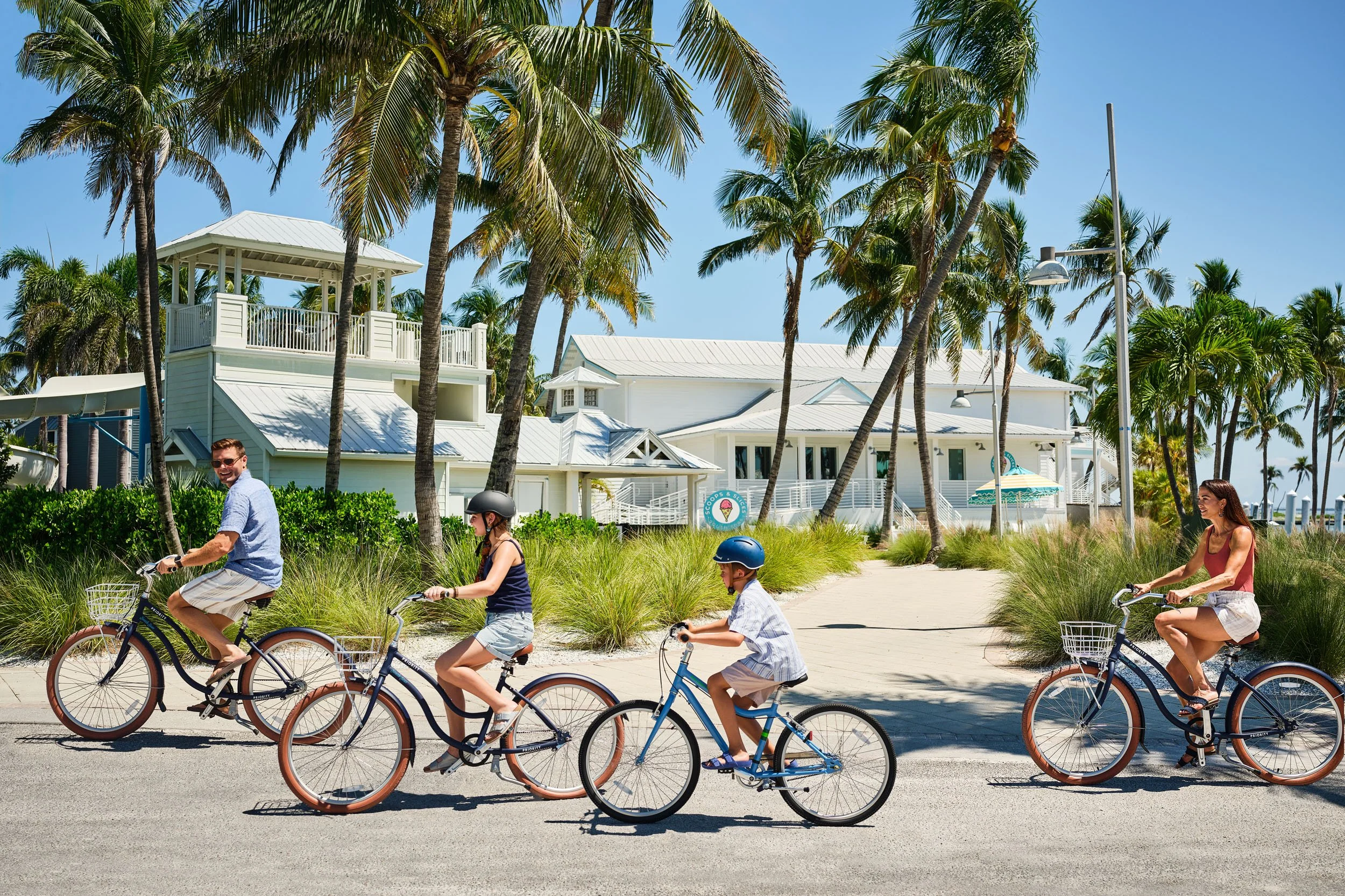 People riding bicycles on a sunny promenade lined with palm trees, with white beach houses in the background, under a clear blue sky.
