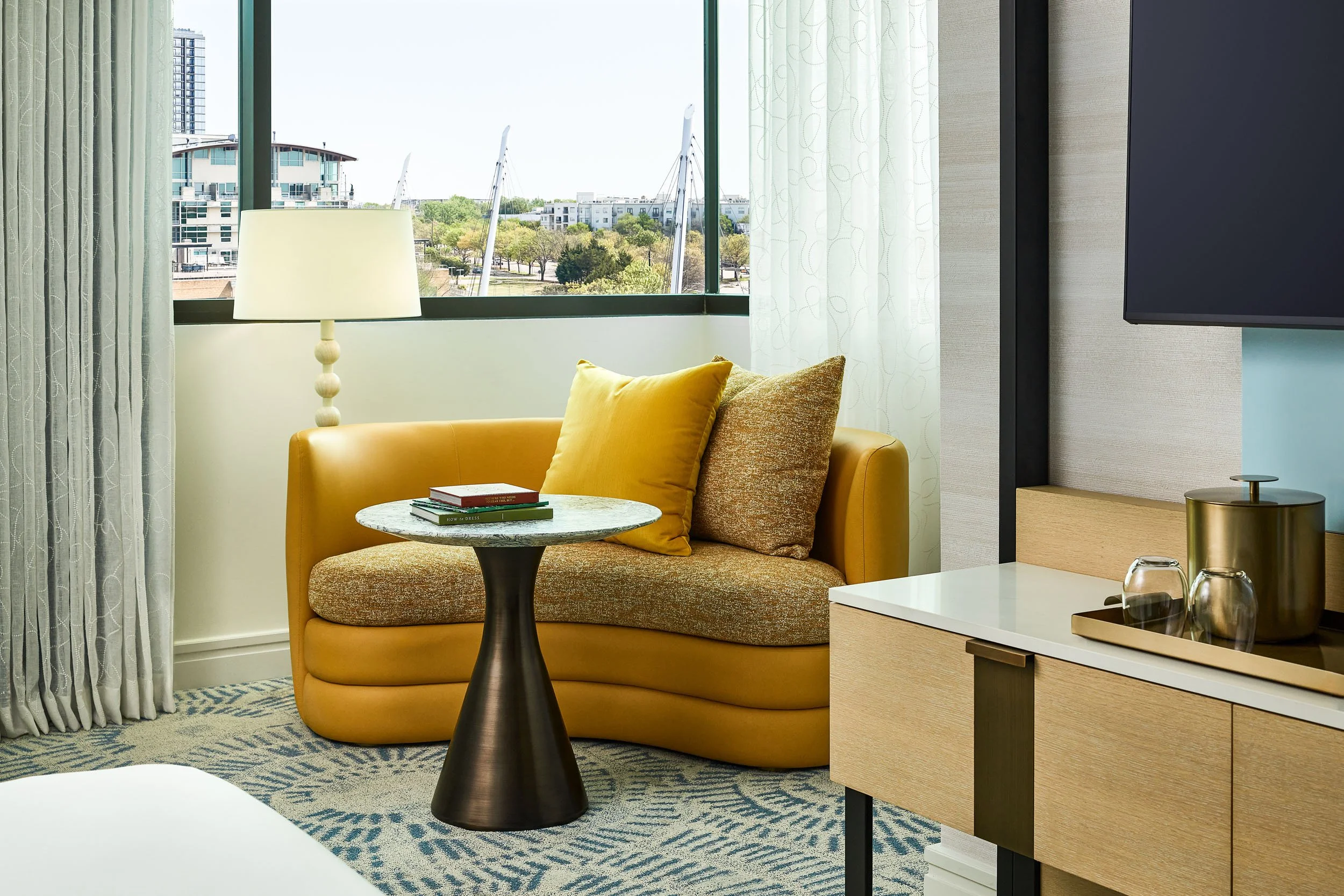 Interior of a modern living room with a yellow sofa, decorative pillows, a round coffee table, a floor lamp, and large windows with curtains, overlooking a cityscape