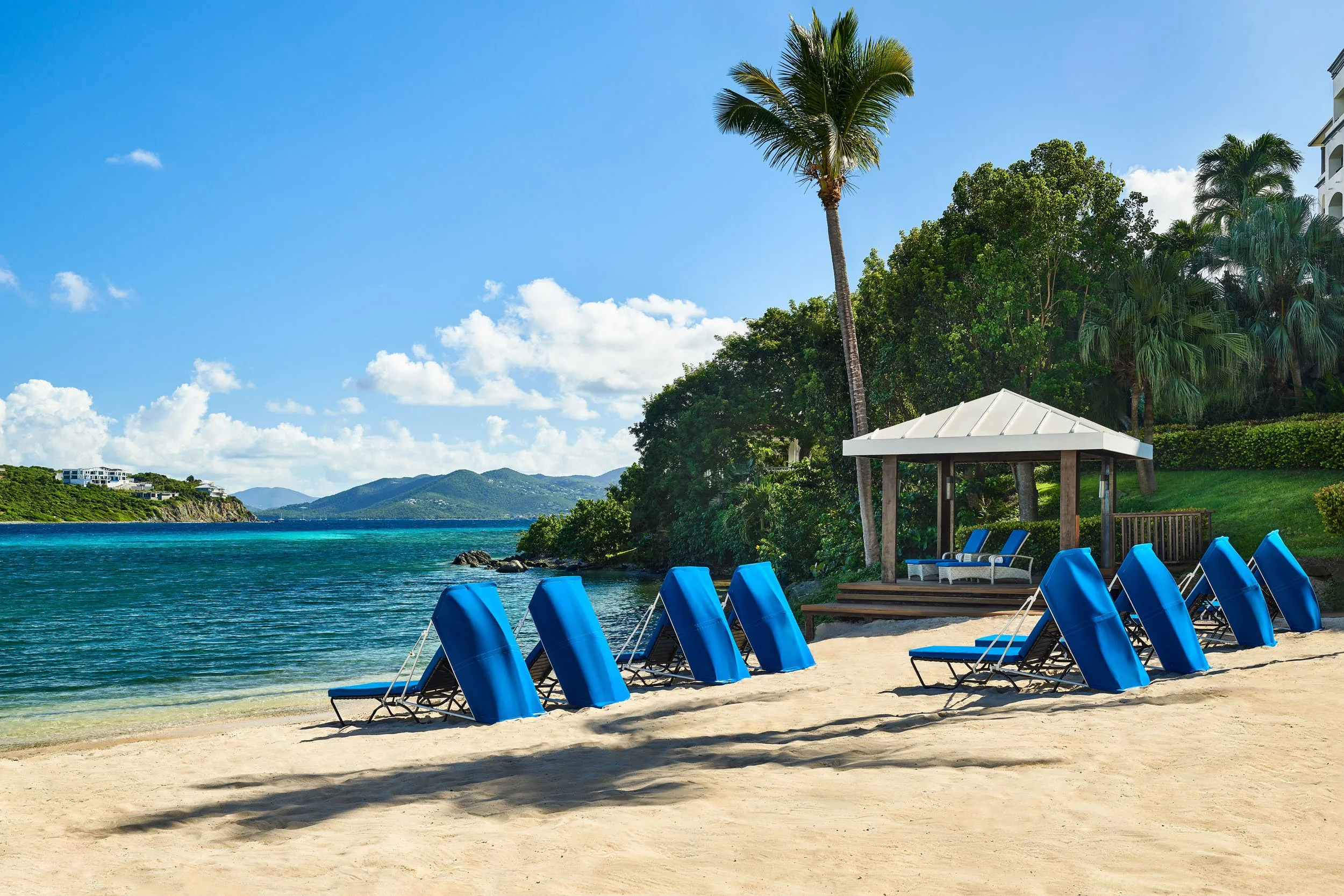 A beach with blue lounge chairs arranged in a row on the sandy shore, a shaded gazebo, green trees, palm trees, clear water, and distant hills under a blue sky with some clouds.