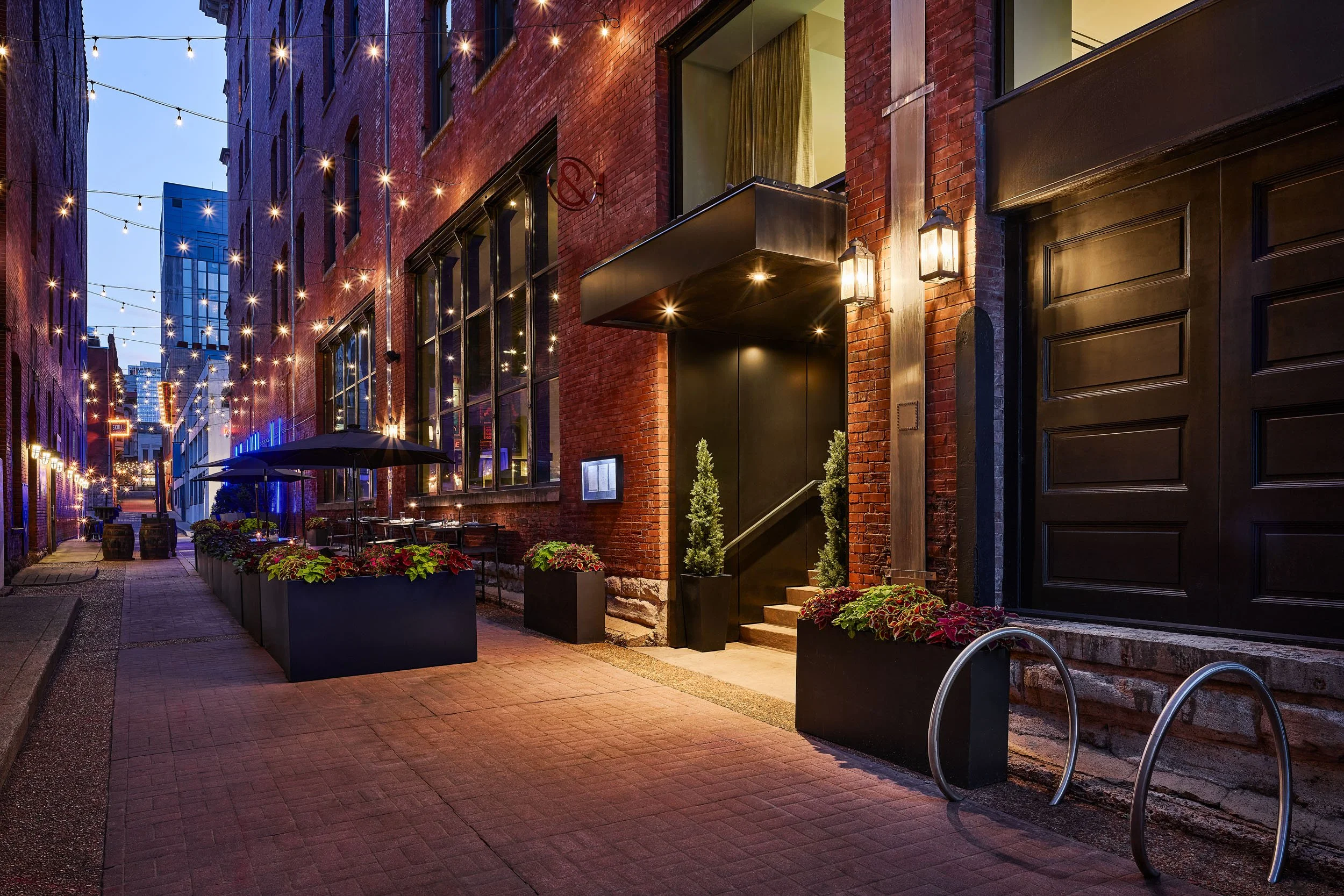 Night scene of a city alleyway with brick buildings, hanging string lights, outdoor seating, potted plants, and a stairway leading to an entrance.