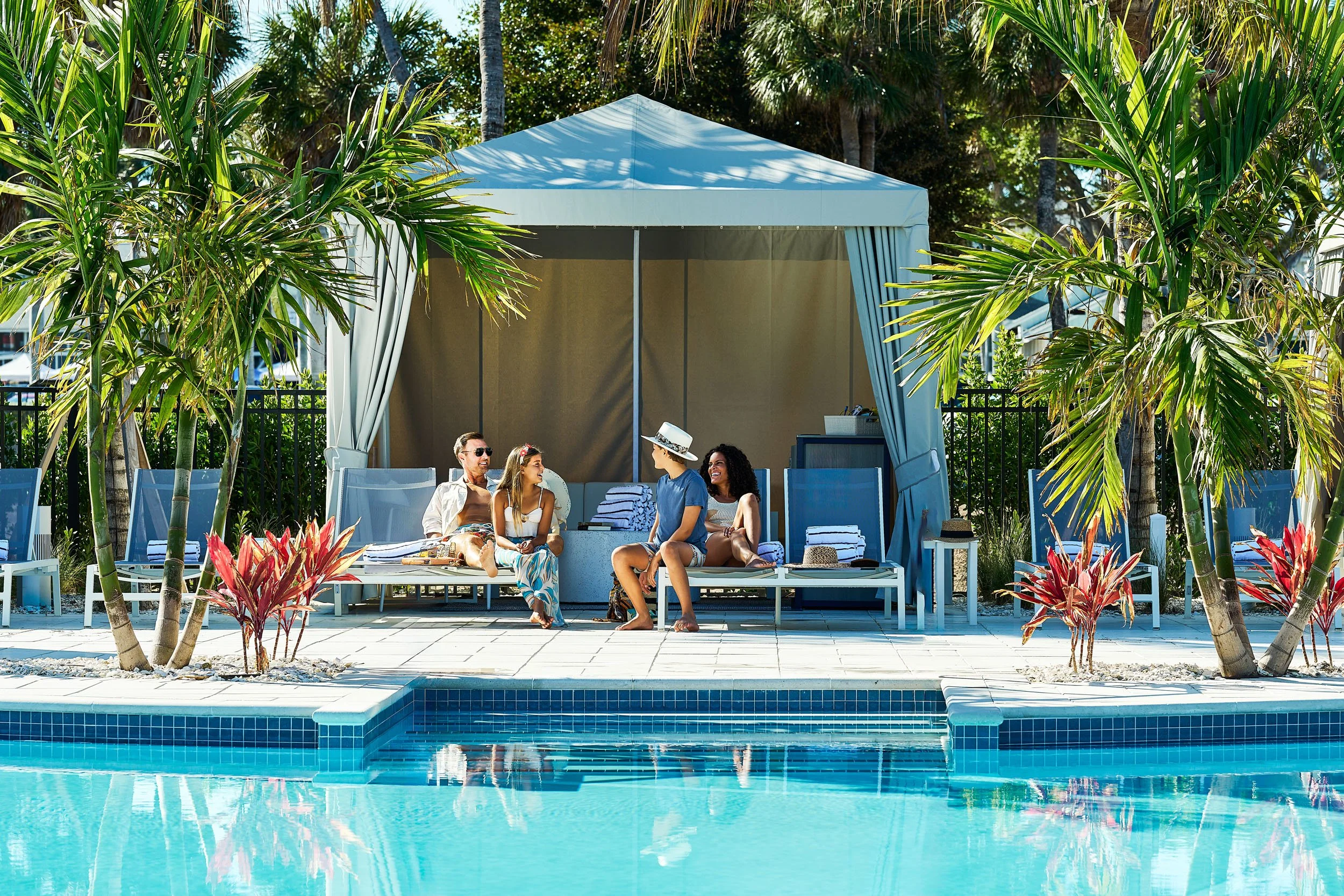 People sitting on poolside lounge chairs under a canopy surrounded by tropical palm trees.