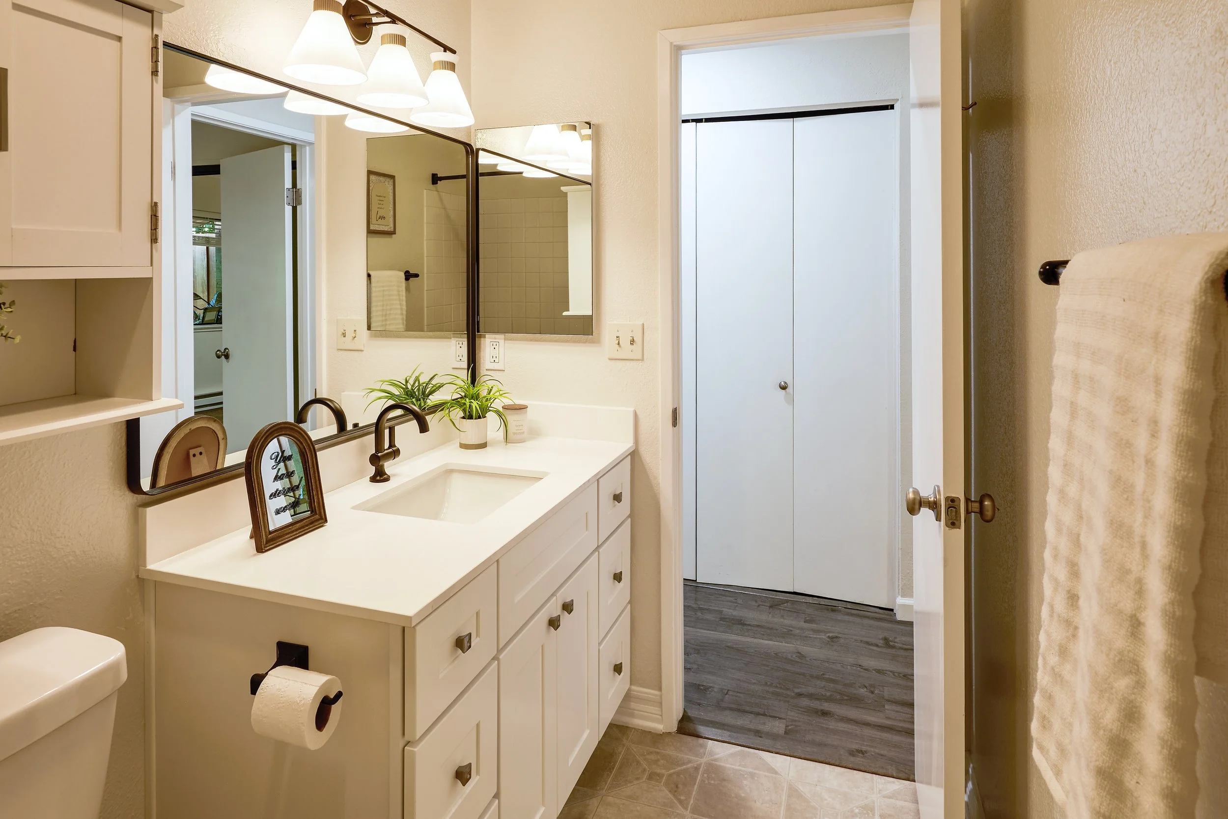 Interior view of a bathroom with a white vanity, black faucets, and a large mirror. There are two small potted plants on the vanity, a framed quote, and a decorative picture frame. The bathroom has beige walls, a towel rack with a towel, and a ceilin