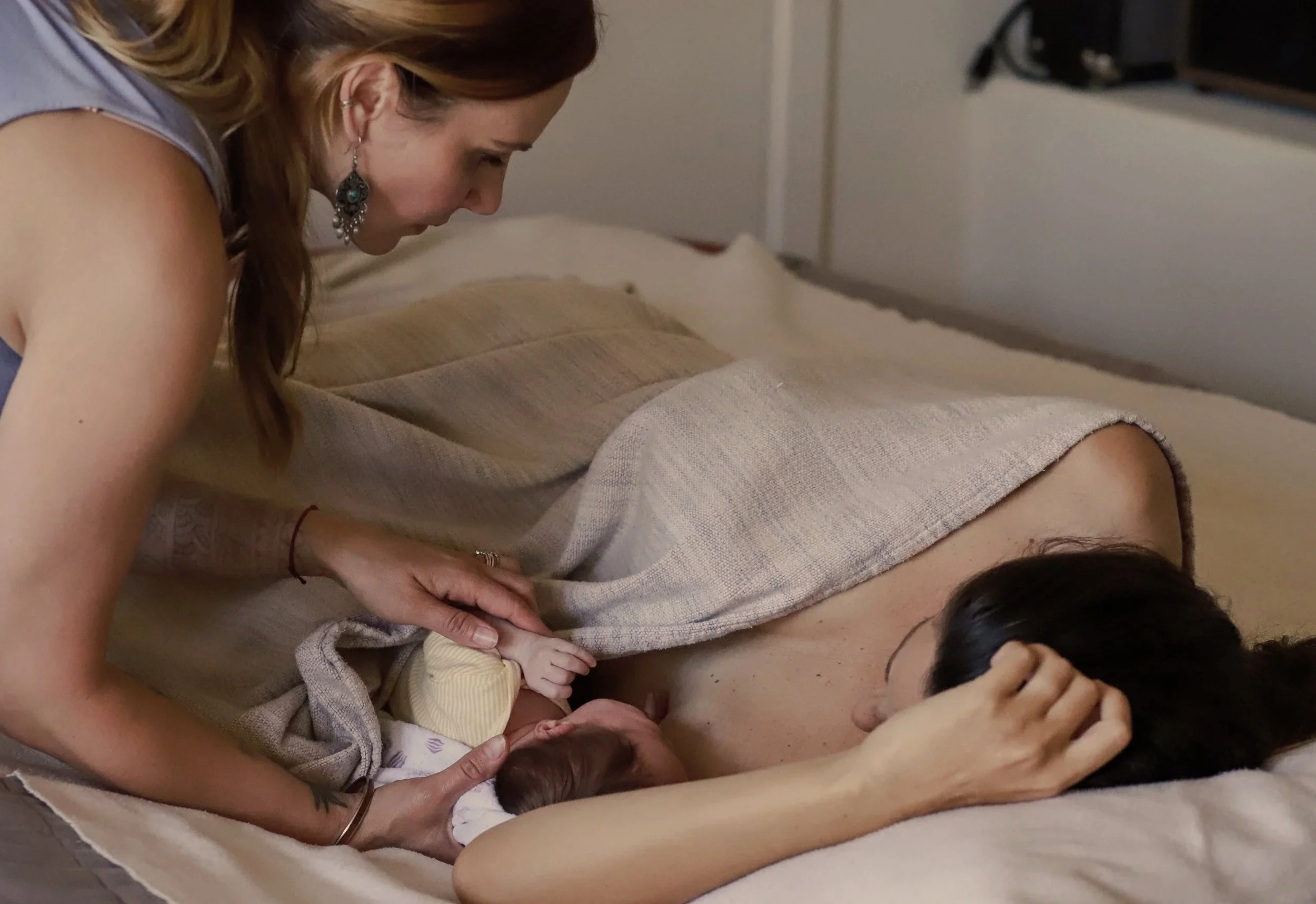 A woman with reddish hair and earrings leans over a person lying on a bed or couch, holding their hand. The person lying down has dark hair and is partially covered by a beige blanket. The scene appears intimate and warm, possibly a comforting or caregiving moment.
