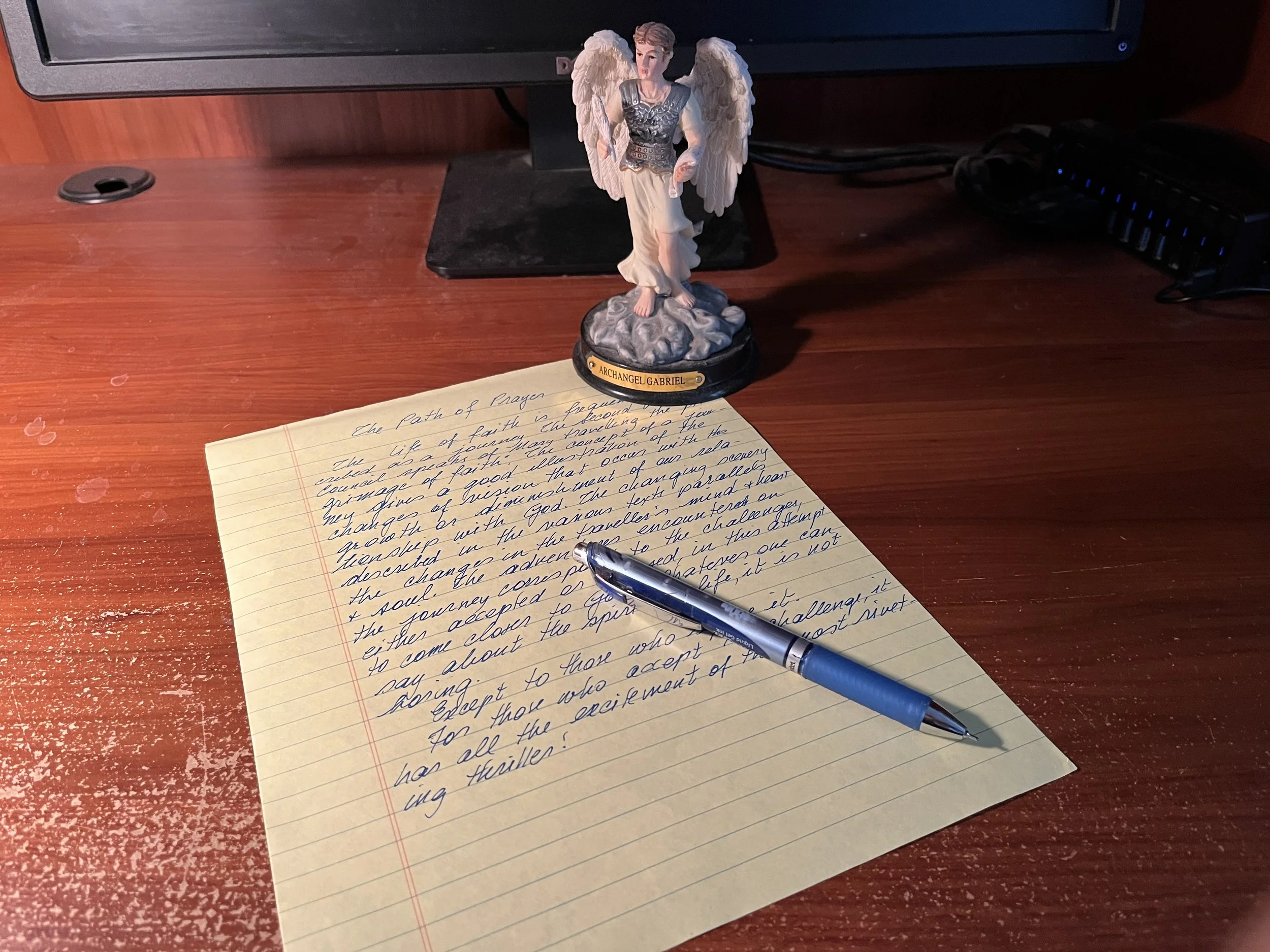A wooden desk with a handwritten sheet of paper, a blue pen resting on it, and a figurine of Archangel Gabriel,.