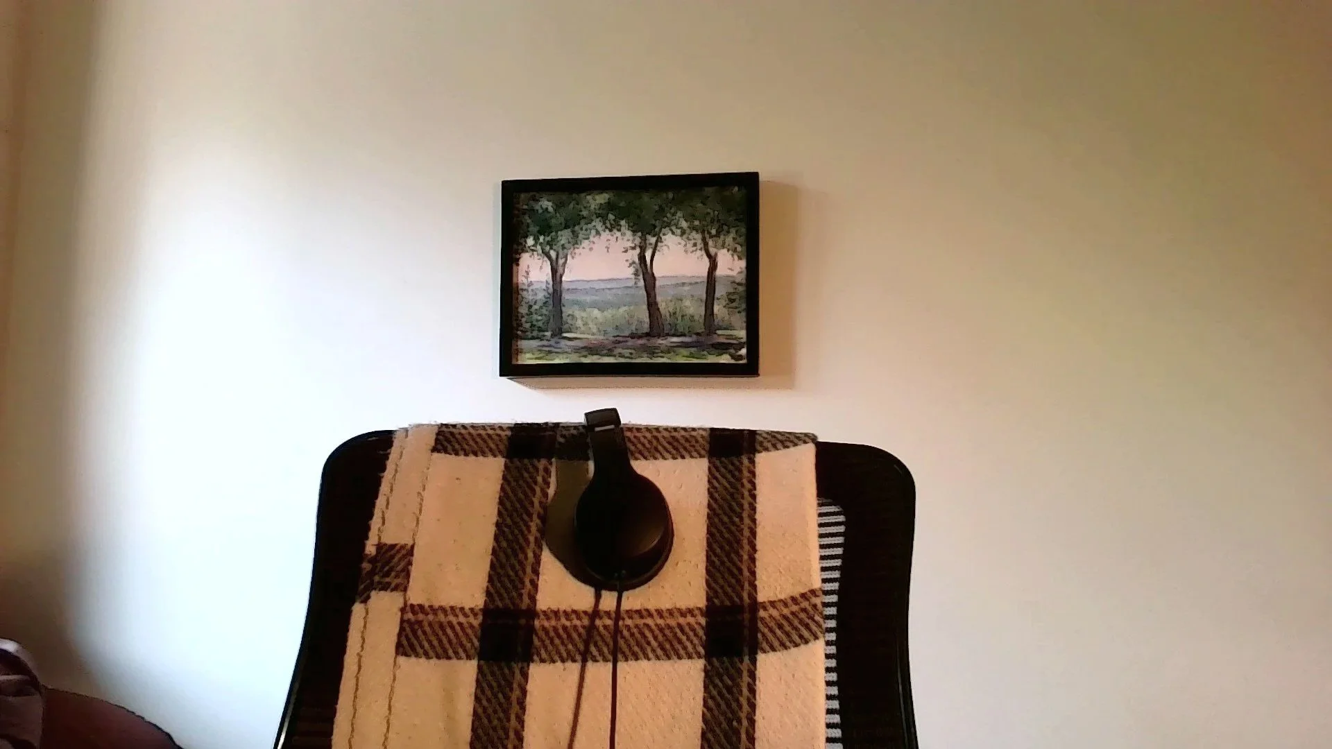 A black chair with a blanket draped over it, a pair of black headphones resting on the cloth, a framed painting of trees hanging in the background.