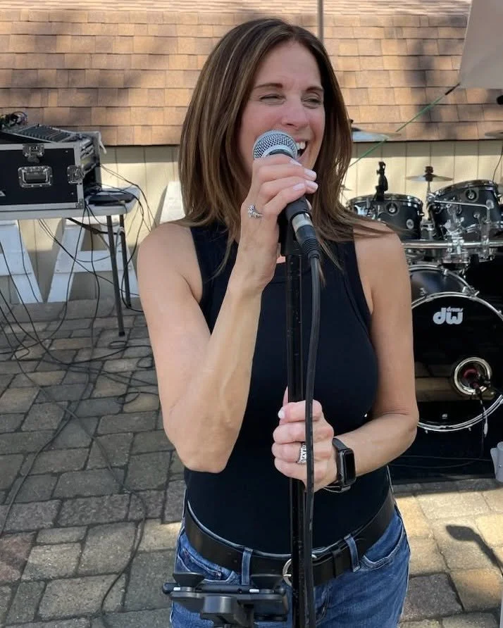 A woman singing into a microphone outdoors with musical equipment behind her.