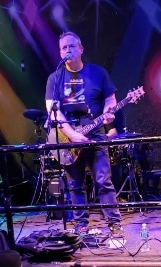 A man performing on stage with an electric guitar surrounded by music equipment, colorful stage lighting, and electronic devices.