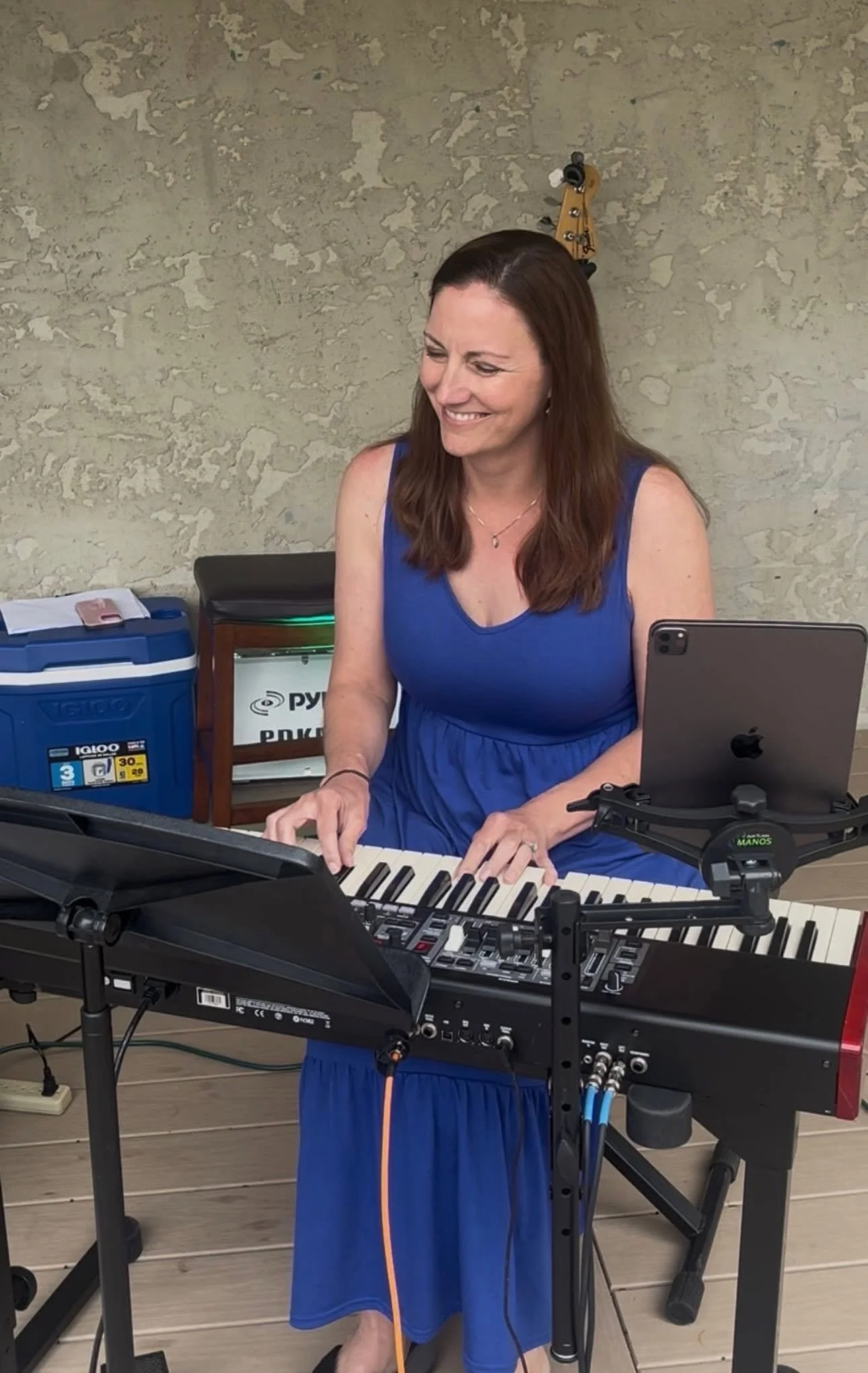 A woman with long brown hair wearing a blue dress playing a keyboard, smiling in a music studio or rehearsal space, with music equipment and a textured wall in the background.