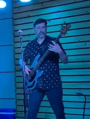 Man playing an electric bass guitar on a stage with wooden panel background.