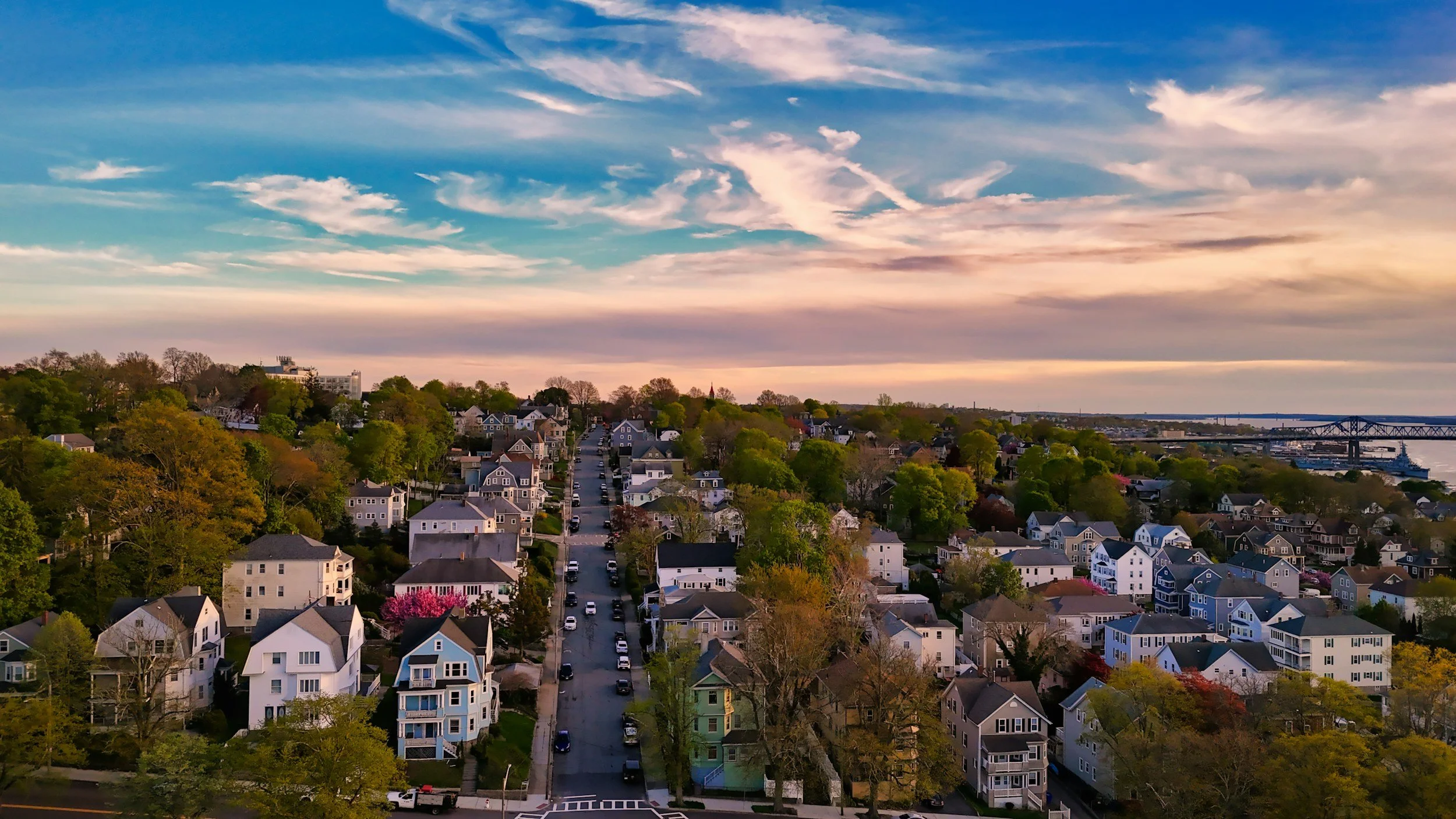 A scenic view of a residential neighborhood with houses of various colors and a river with a bridge in the background, under a partly cloudy sky during sunset.