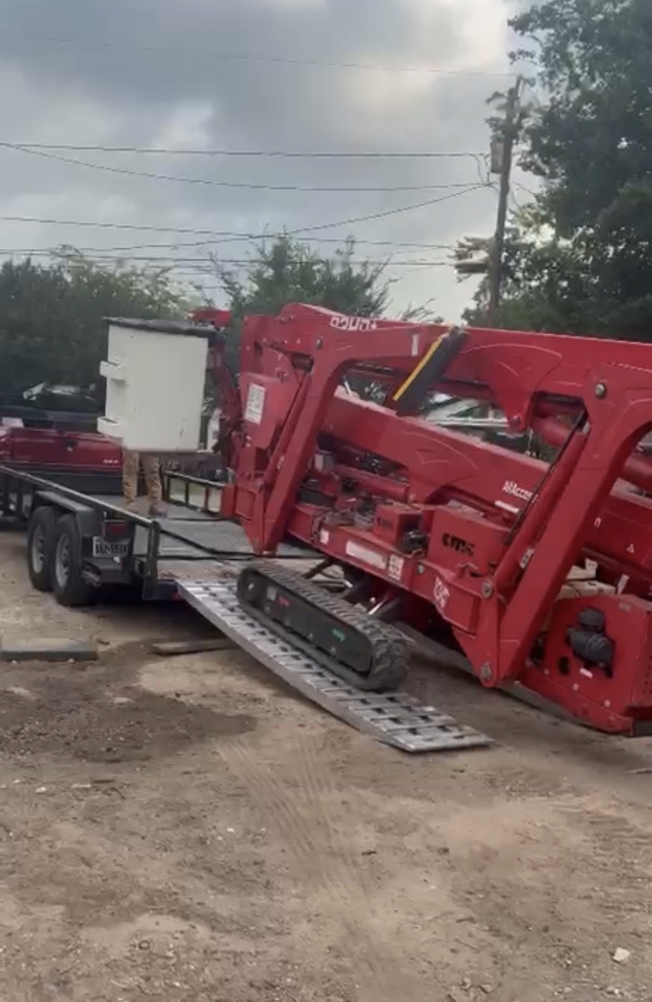 A red crane on a flatbed trailer parked on a dirt surface, with power lines, trees, and a cloudy sky in the background.