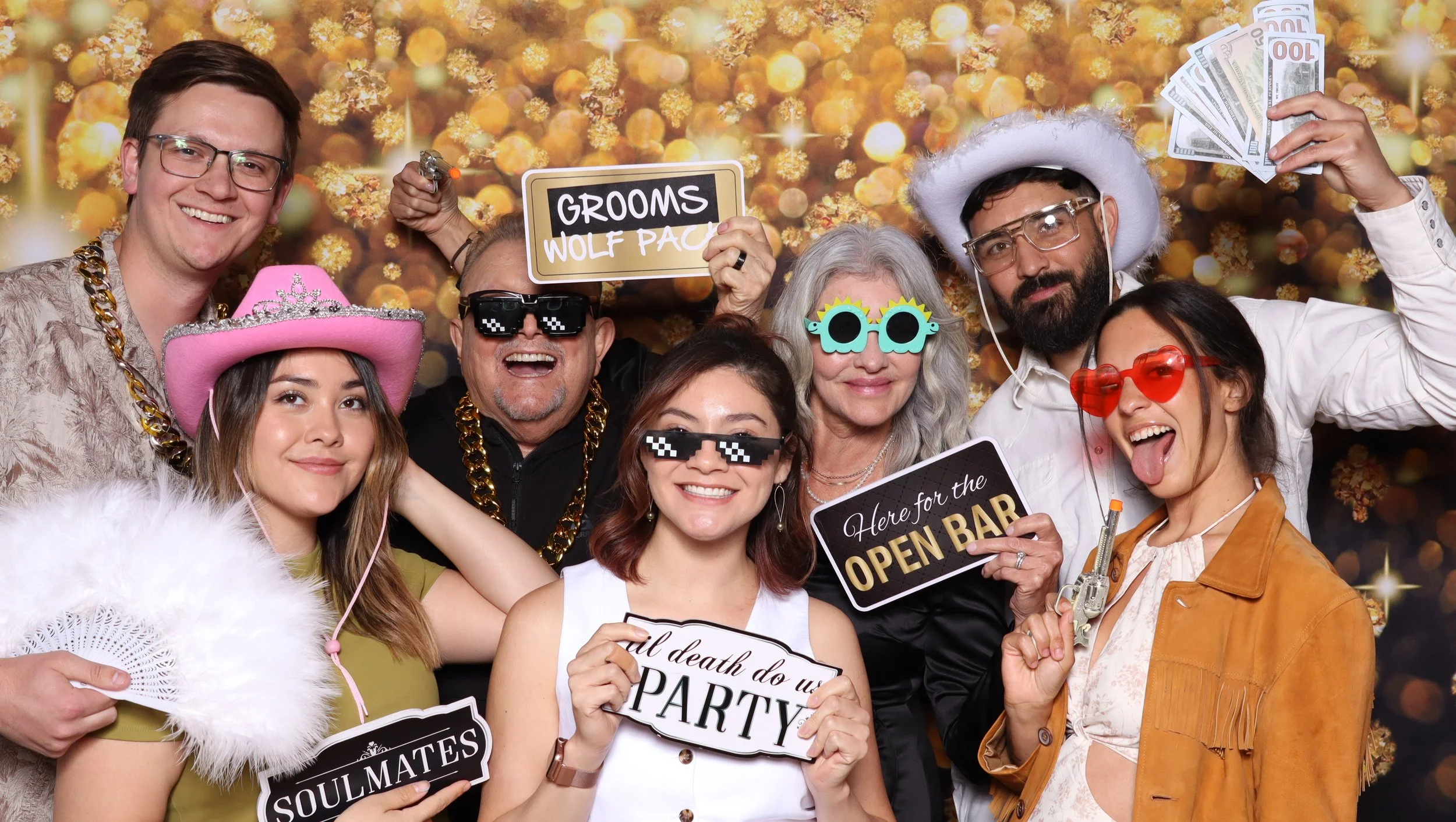 Group of guests posing with signs and accessories in front of a gold photo booth backdrop.
