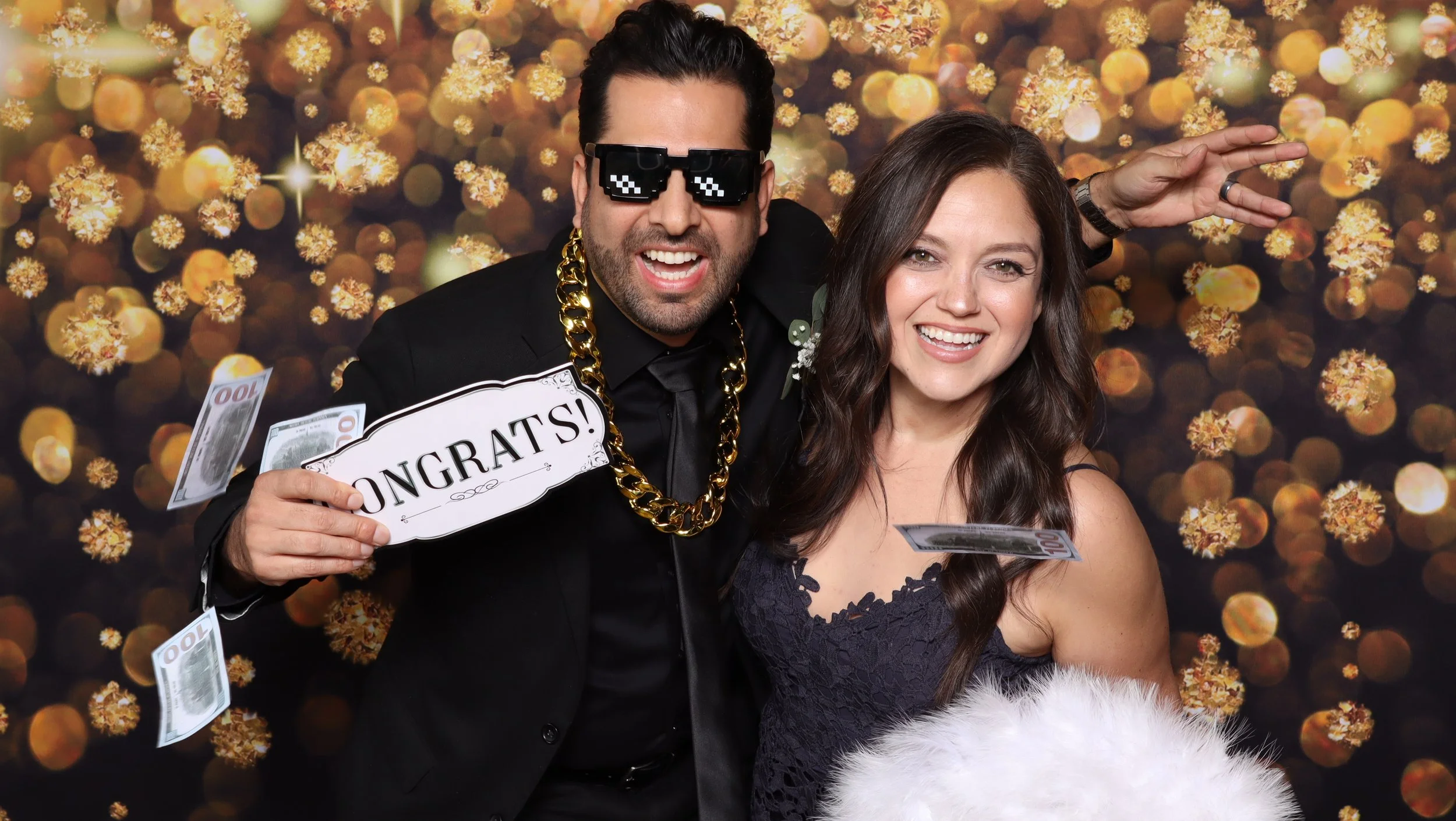 Two guests posing with photo booth accessories in front of a gold backdrop.