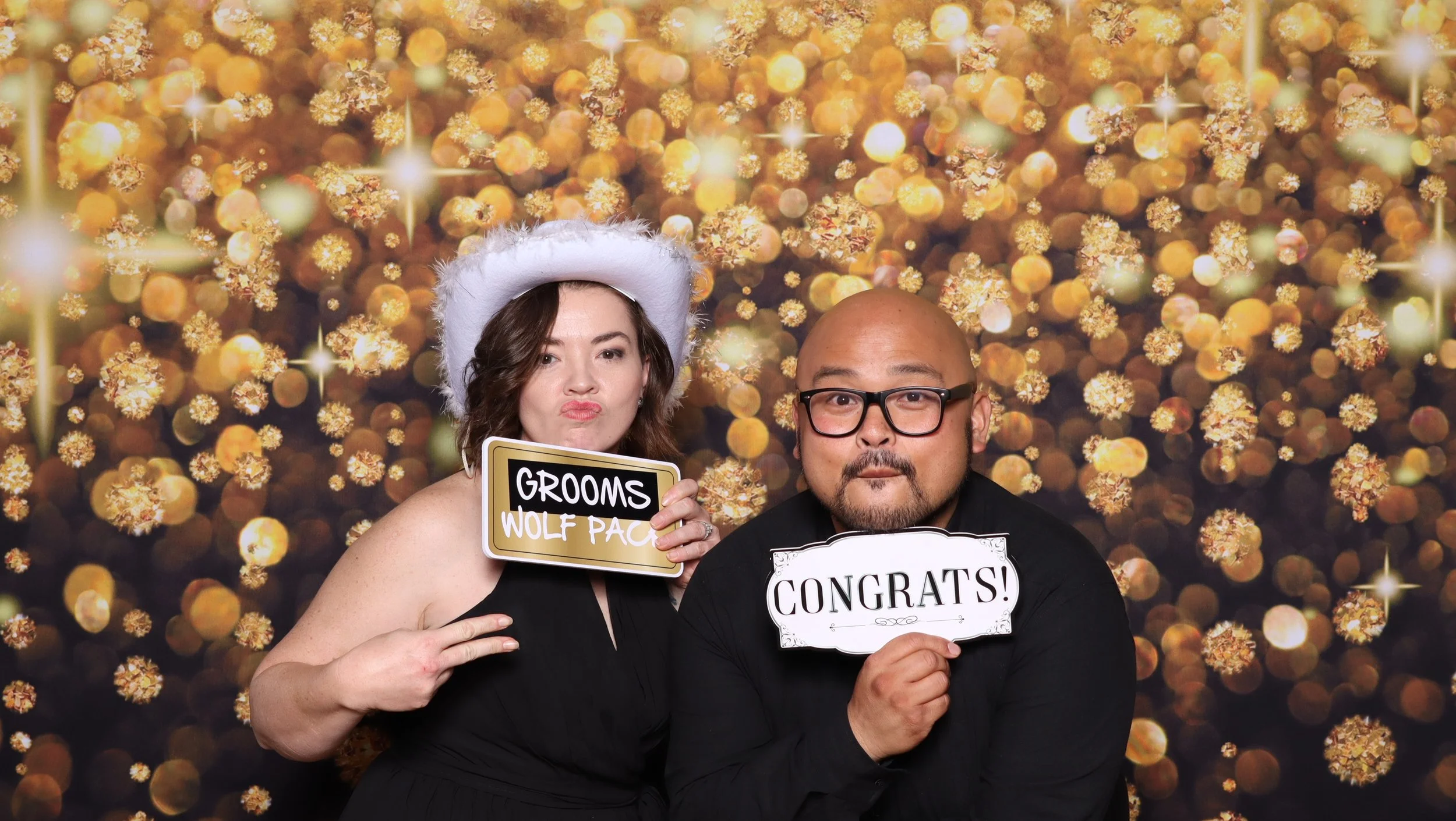 Two wedding guests posing with signs in front of a gold photo booth backdrop.