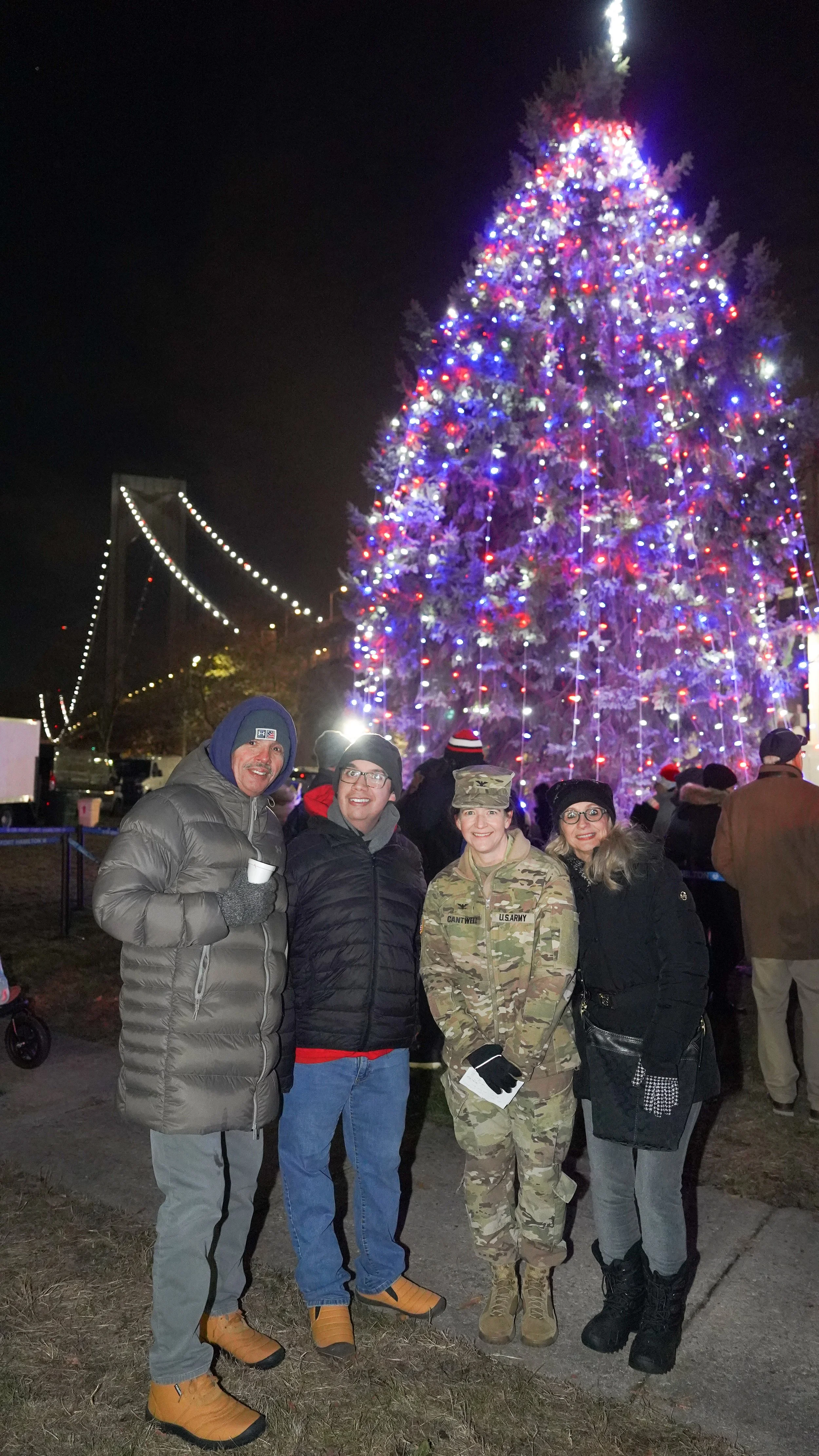 Four people standing in front of a large Christmas tree decorated with colorful lights at night. The group includes a man in a gray puffer jacket, a man in a black jacket, a woman in a U.S. Army uniform, and a woman in a black coat and glasses. The background features a nighttime scene with a decorated bridge and other people celebrating.