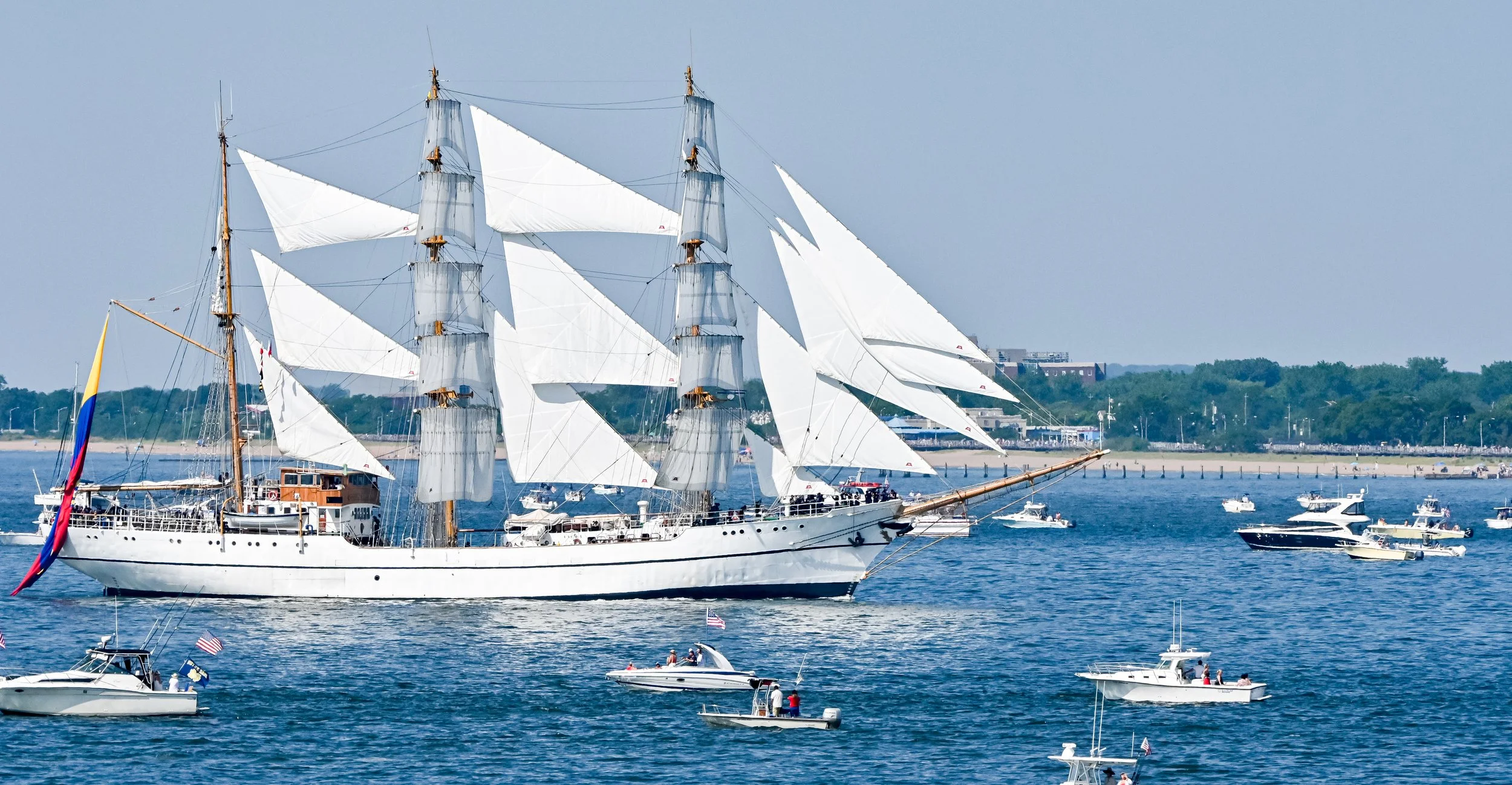 A large white sailing ship with multiple masts and white sails sailing on a body of water with smaller boats and yachts around it.