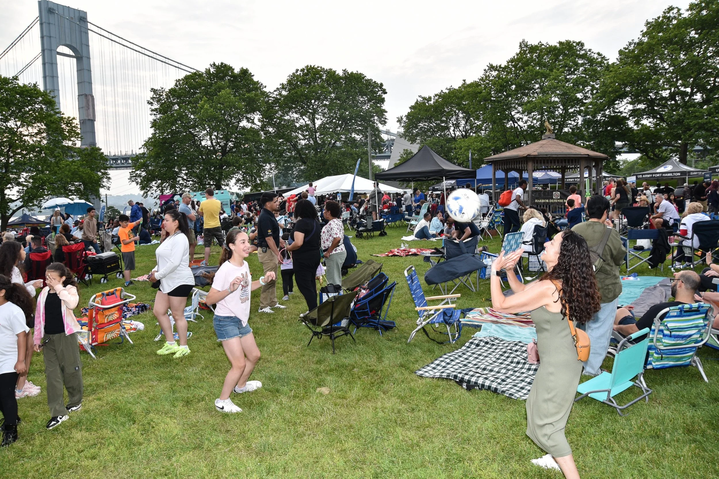 People gathered at an outdoor event with tents, chairs, and trees. The event is near a bridge under a cloudy sky.