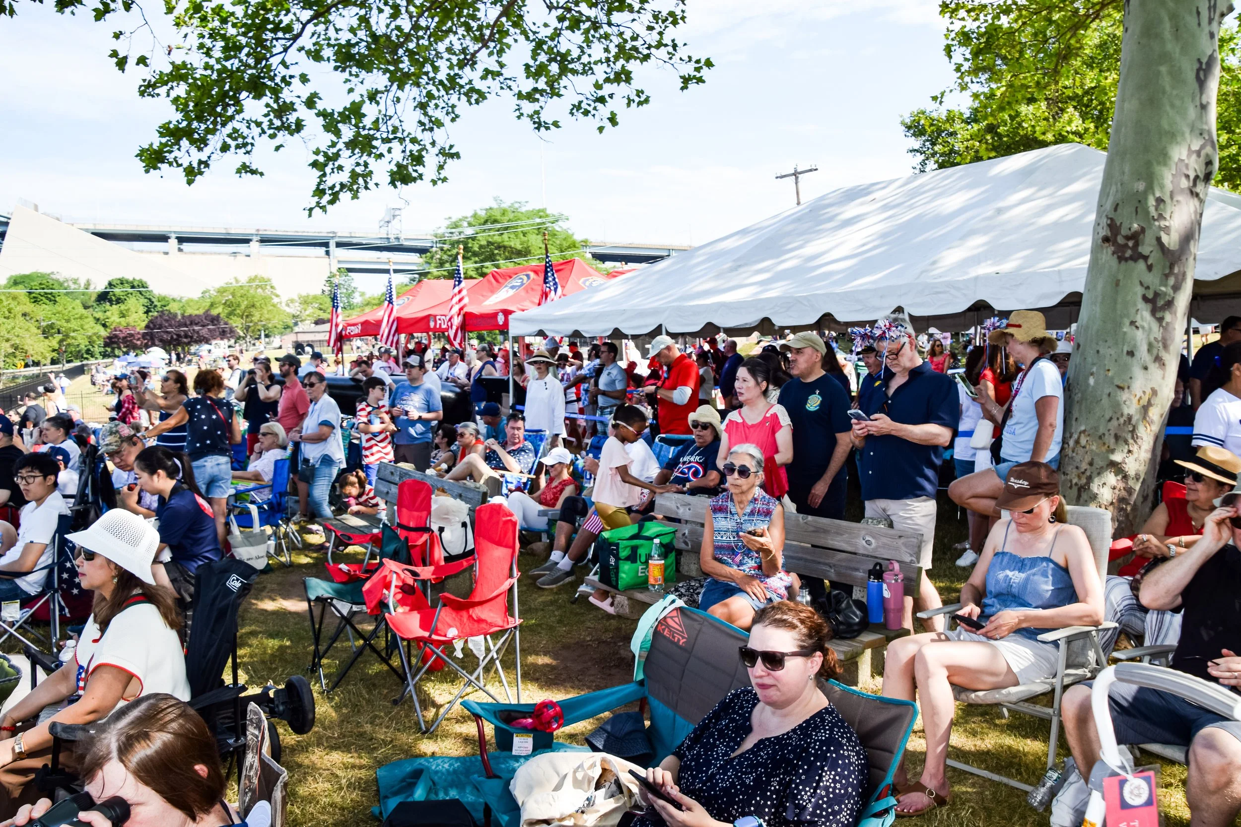 Crowd gathered outdoors at a patriotic event with red, white, and blue tents, American flags, and people sitting and standing under trees on a sunny day.