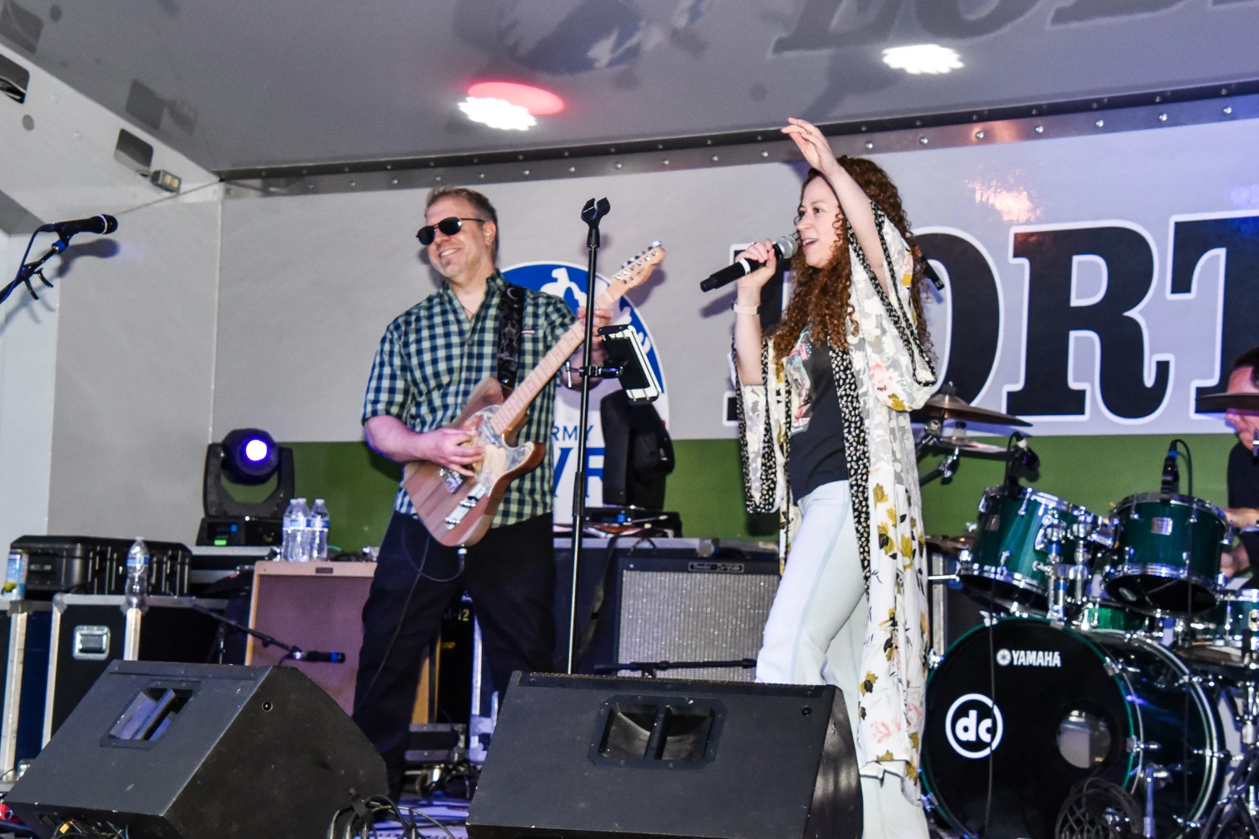 Musicians performing on stage with guitars and drums, a man wearing sunglasses and a checkered shirt playing guitar, a woman with curly hair holding a microphone, singing, with stage equipment and water bottles in front of them.